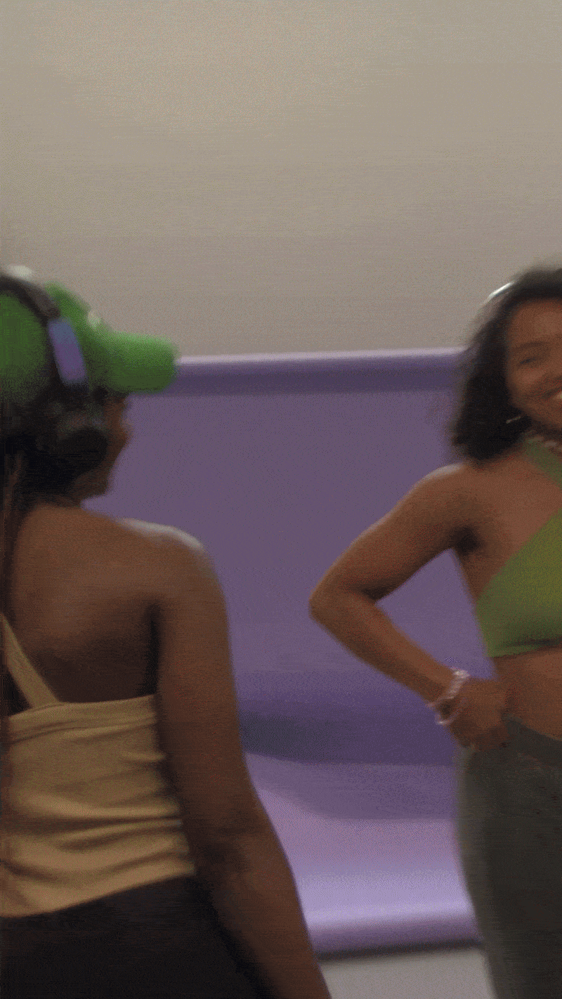 Two women standing indoors, one wearing a beige top and the other in a green top, smiling and chatting, with the woman on the left wearing a green hairdryer on her head.