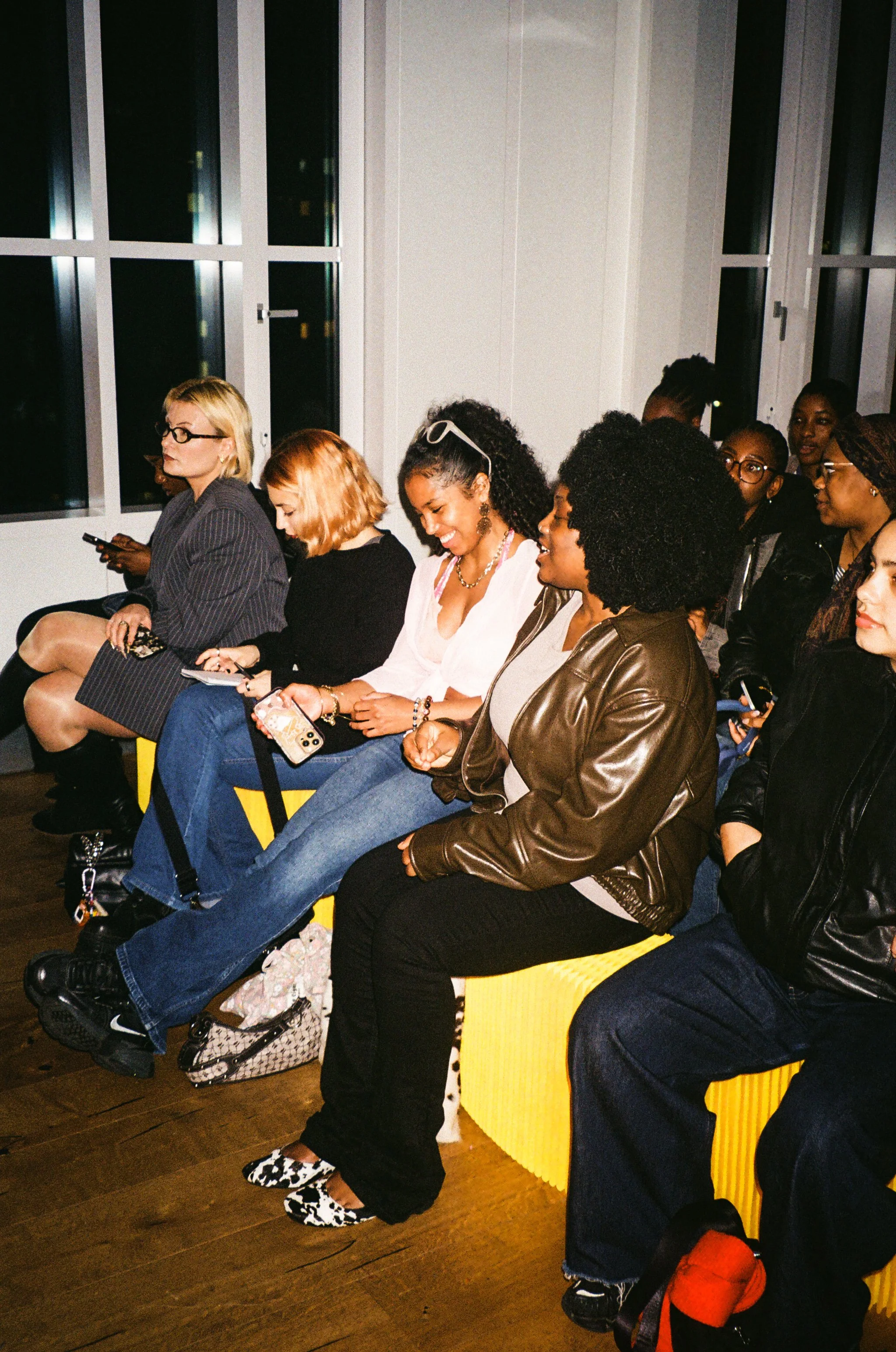 Group of women seated on yellow benches at an indoor event, some looking at their phones, with large windows in the background.