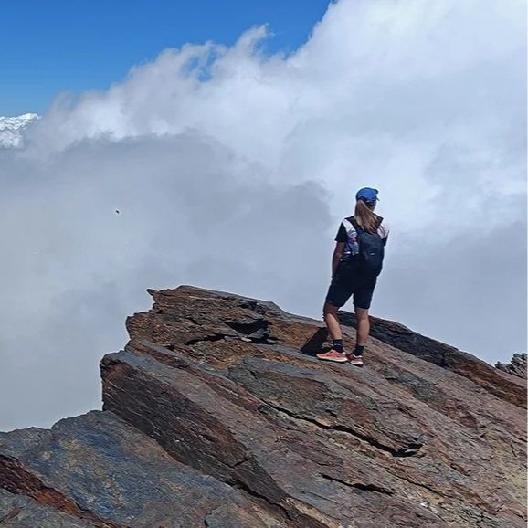 A woman wearing a hiking back pack stands at the edge of a cliff surrounded by white clouds.