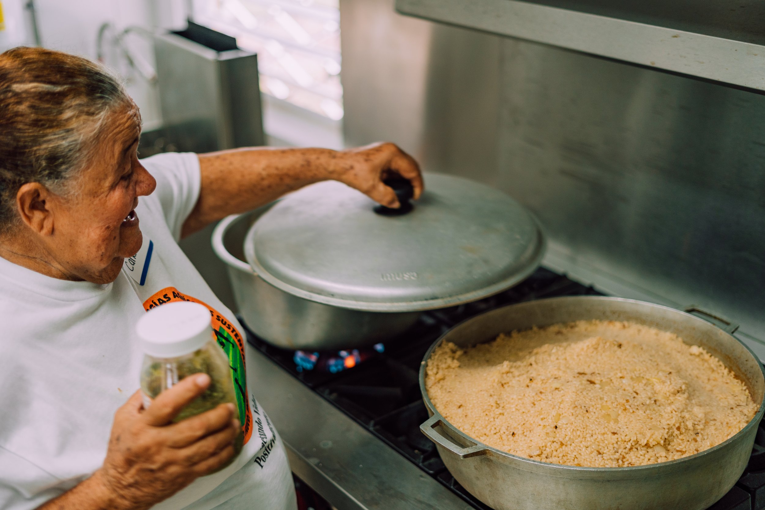 An elderly woman cooking on a stove, stirring a large pot of cornbread crumbs while holding a spice jar in her hand.