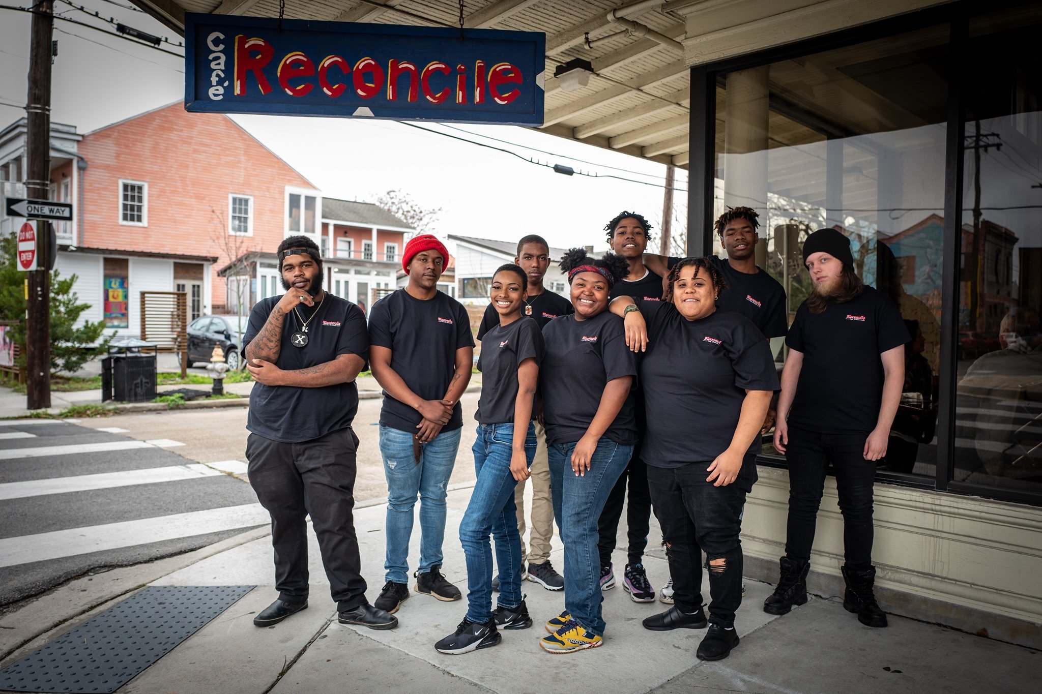 A group of young people stand outside of a restaurant on the sidewalk in front of a sign that reads, "Cafe Reconcile."