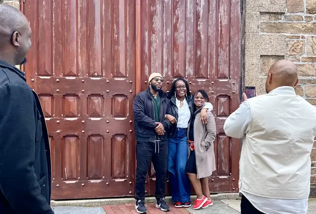 Three people posing for a photo in front of a large wooden door, with two people taking the picture.