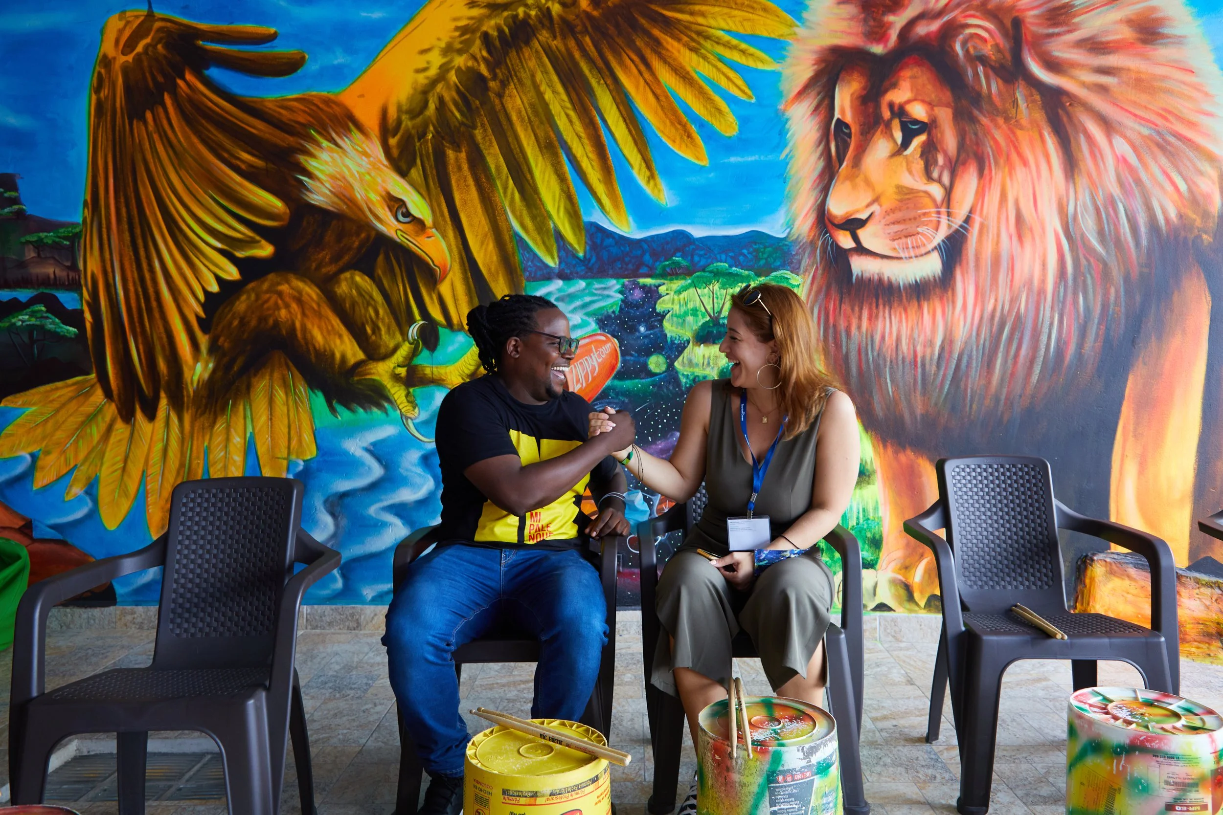 Two people sitting and smiling while engaging in a friendly arm wrestling pose in front of a colorful wall mural depicting a large lion and an eagle in a lush, vibrant landscape.