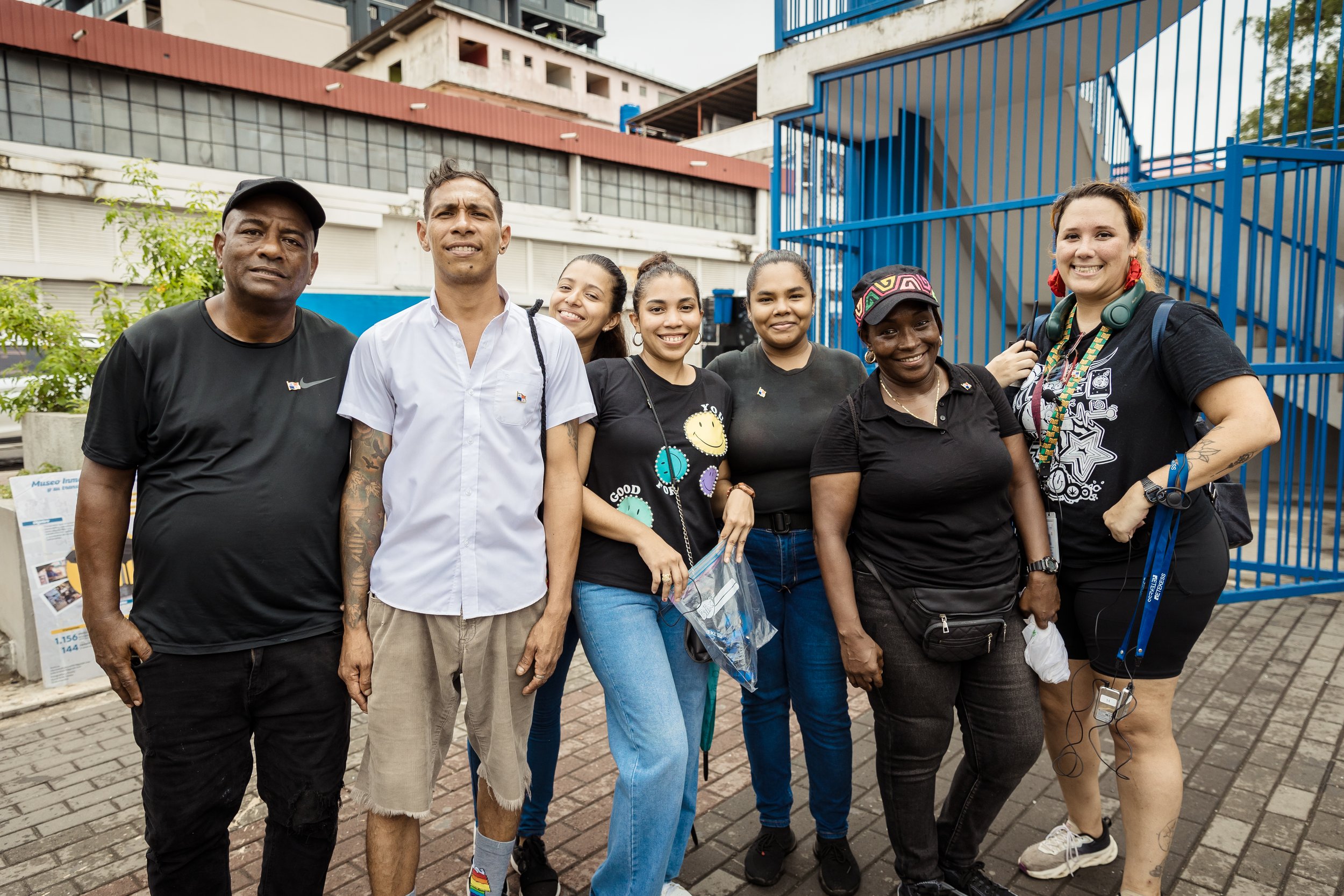 An elderly woman cooking on a stove, stirring a large pot of cornbread crumbs while holding a spice jar in her hand.