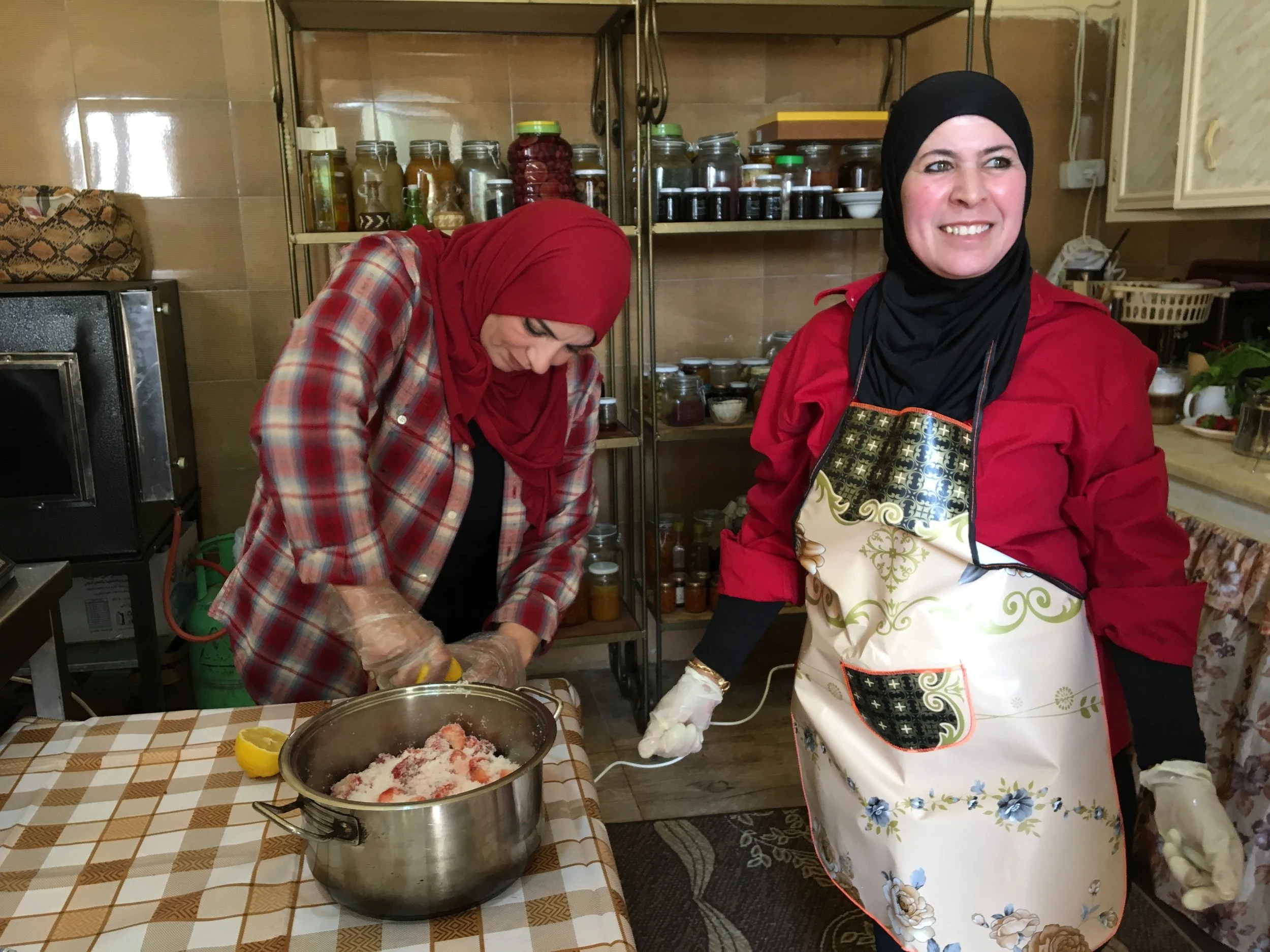 Two women in a kitchen, one wearing a red headscarf and plaid shirt chopping vegetables, and the other wearing a black headscarf, red jacket, and floral apron, smiling.