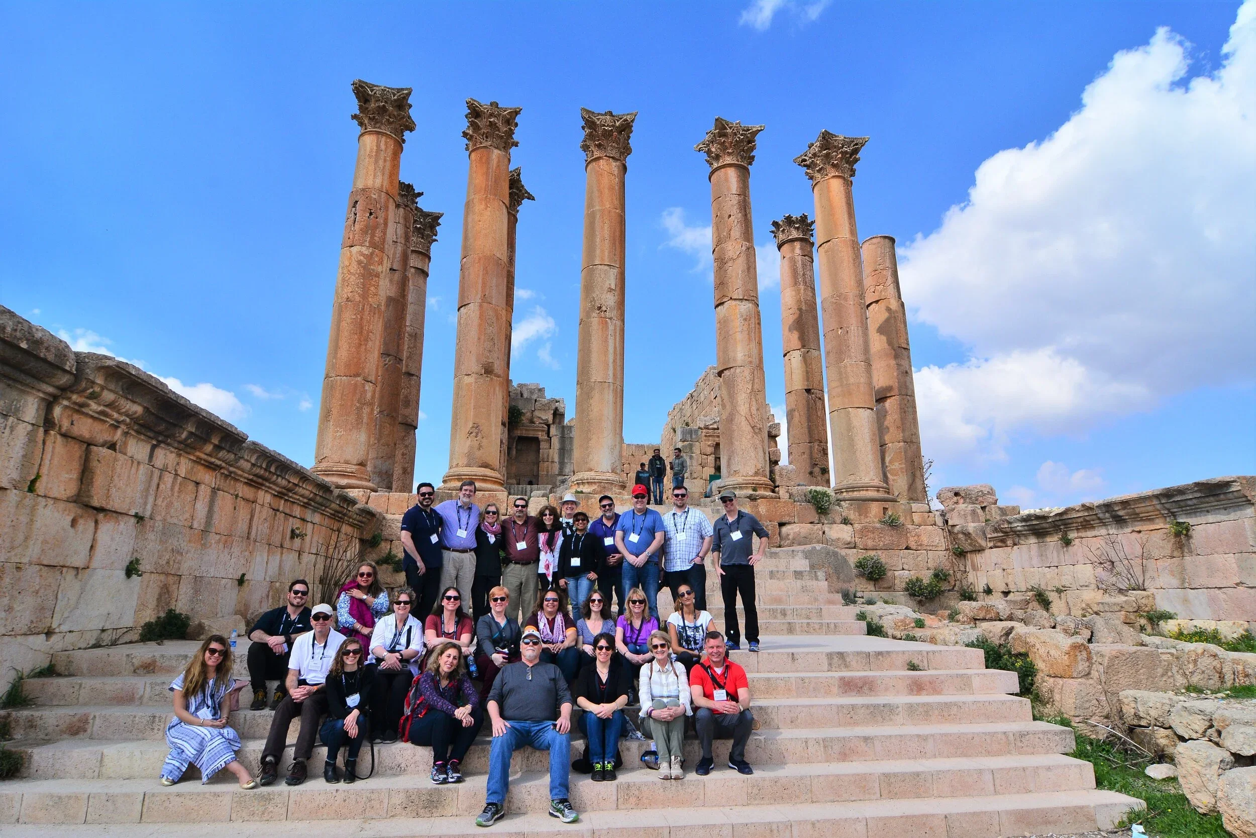 A group of tourists poses on stone steps in front of ancient Roman columns at a historical site under a partly cloudy blue sky.