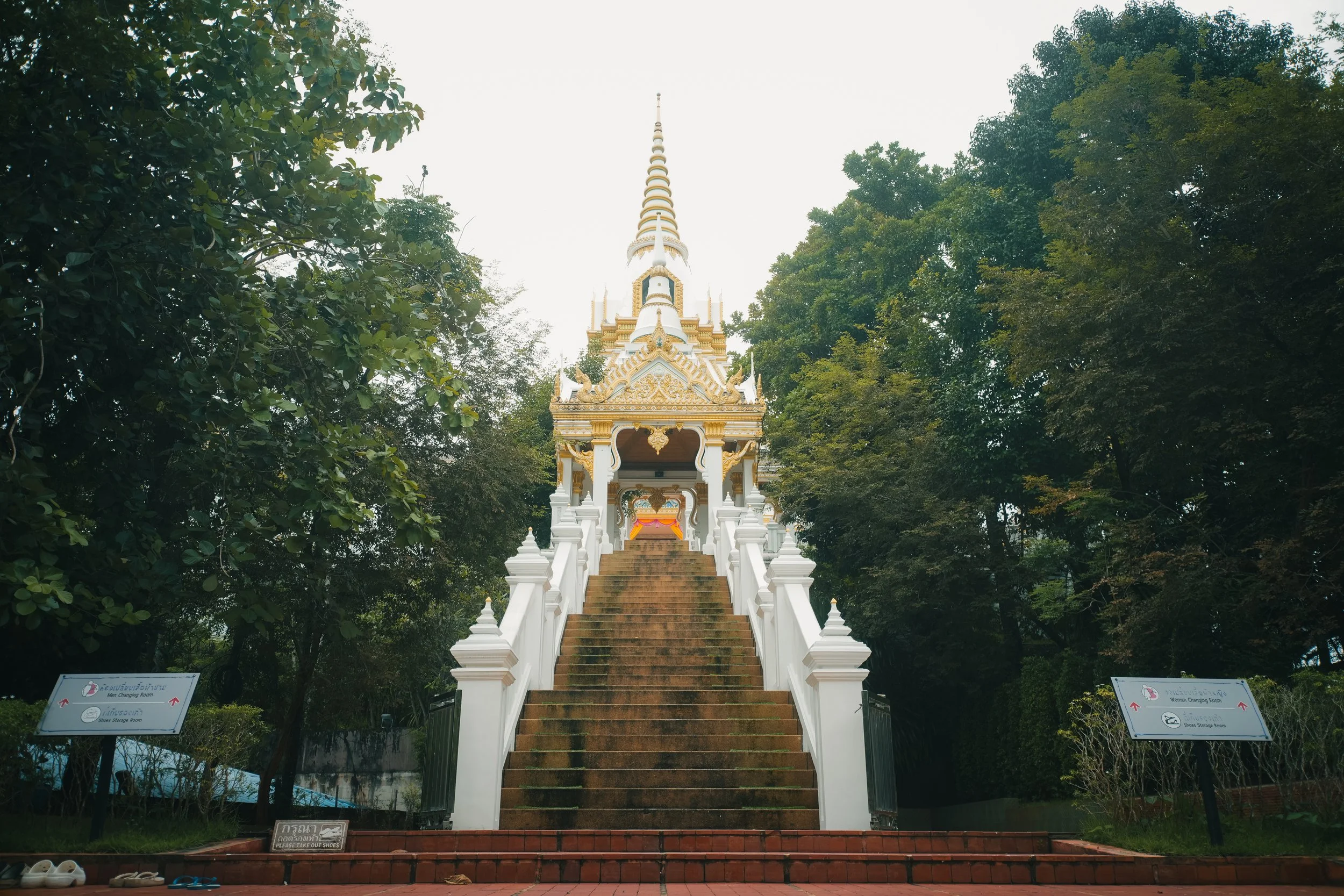 A white and gold Thai-style temple with a high spire, accessed by brown stairs flanked by white railings, surrounded by green trees.