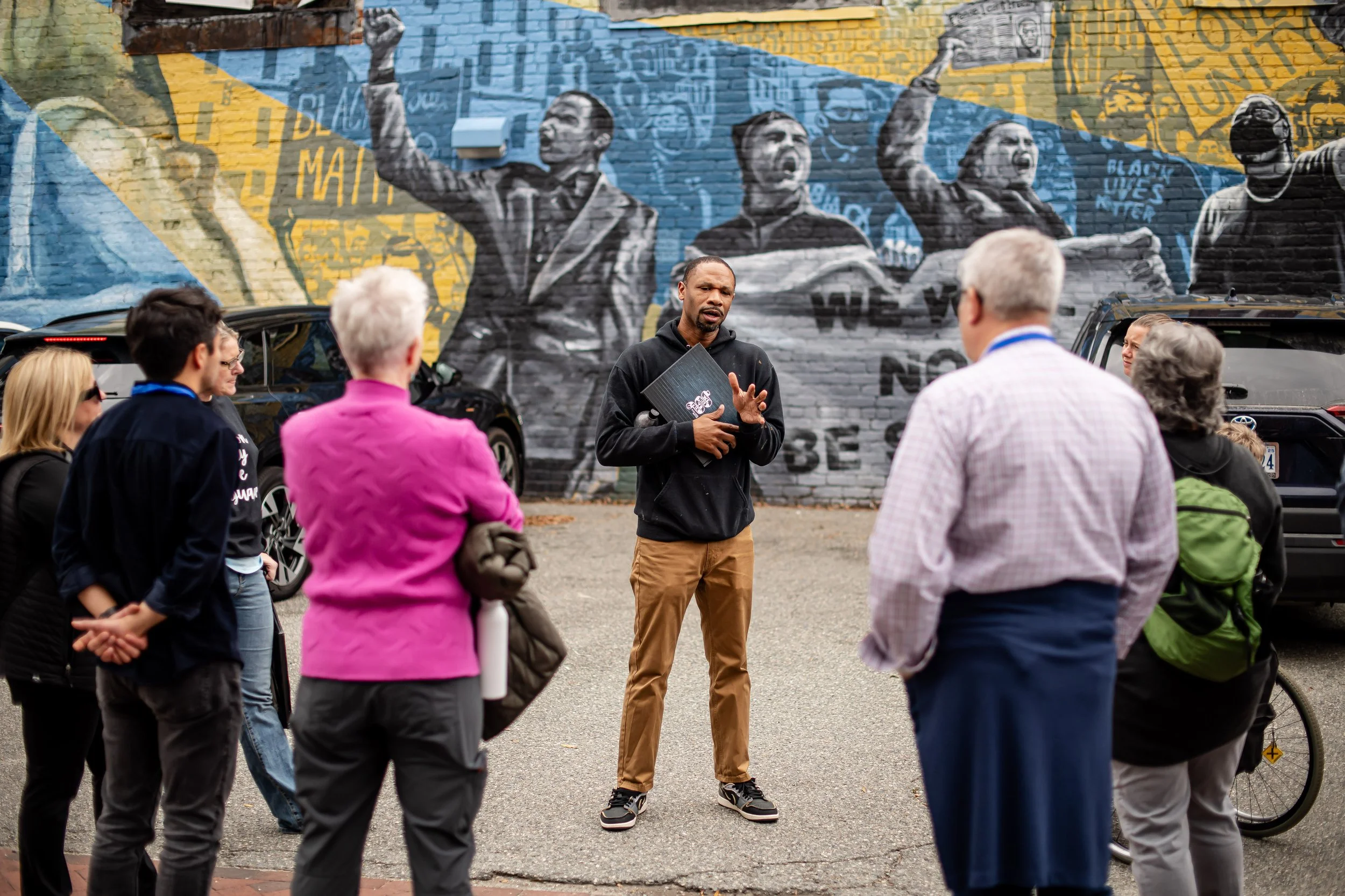 A man giving a speech or tour to a group of people outdoors, with a mural of protesters and messages in the background.