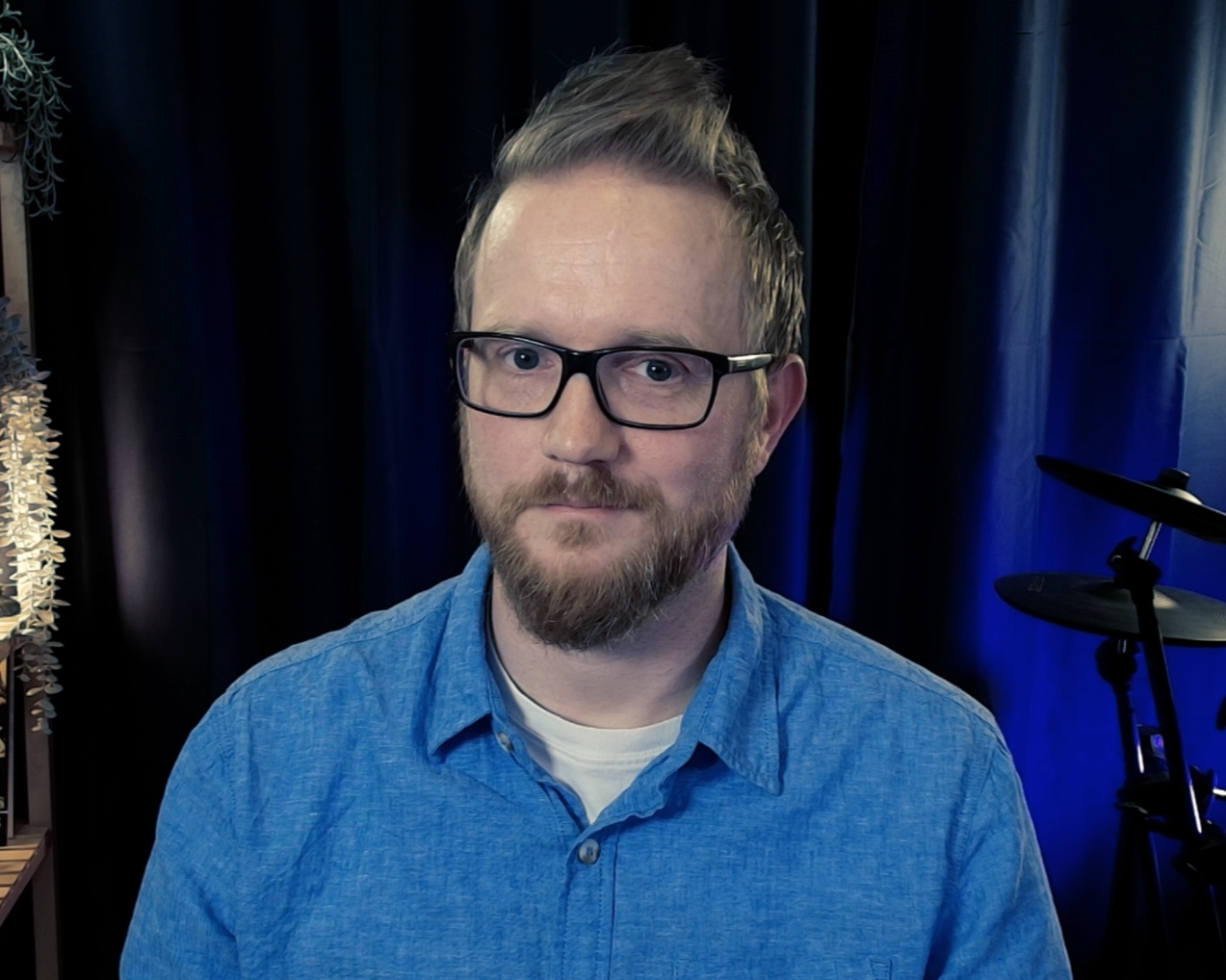 A man with glasses, a beard, and short hair, wearing a blue button-up shirt, sitting in front of a dark curtain background, with a drum set visible on the right side.