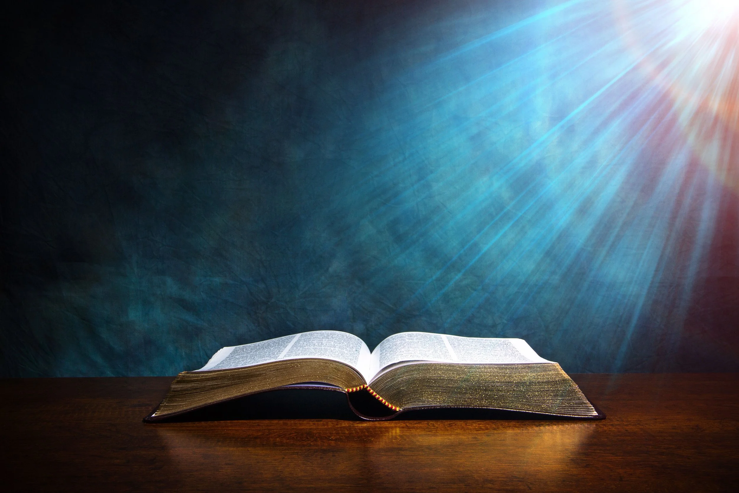 An open Bible on a wooden surface with a dark background and rays of light shining from the top right.