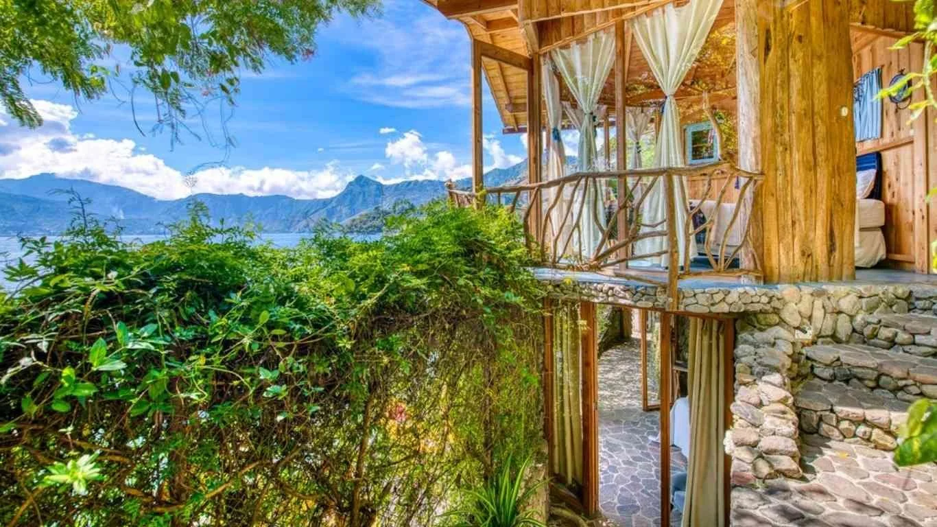 A wooden balcony attached to a rustic cabin with a view of mountains and a lake in the background, surrounded by greenery and blue skies.