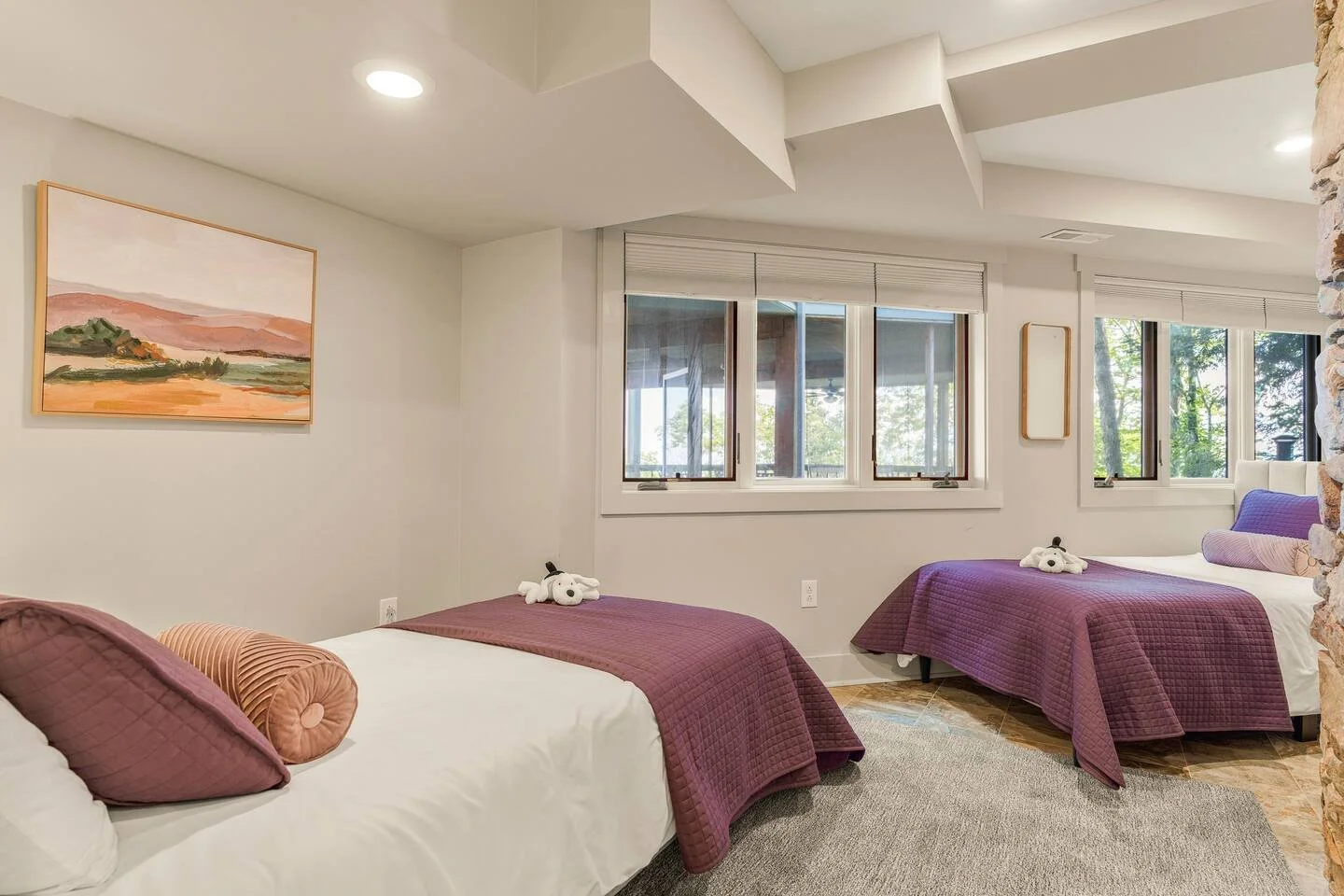 A bedroom with two twin beds covered with purple quilts, each with plush dog toys on top, pillows, and a rug on the floor. There is a window with a view of trees outside, and a painting on the wall.