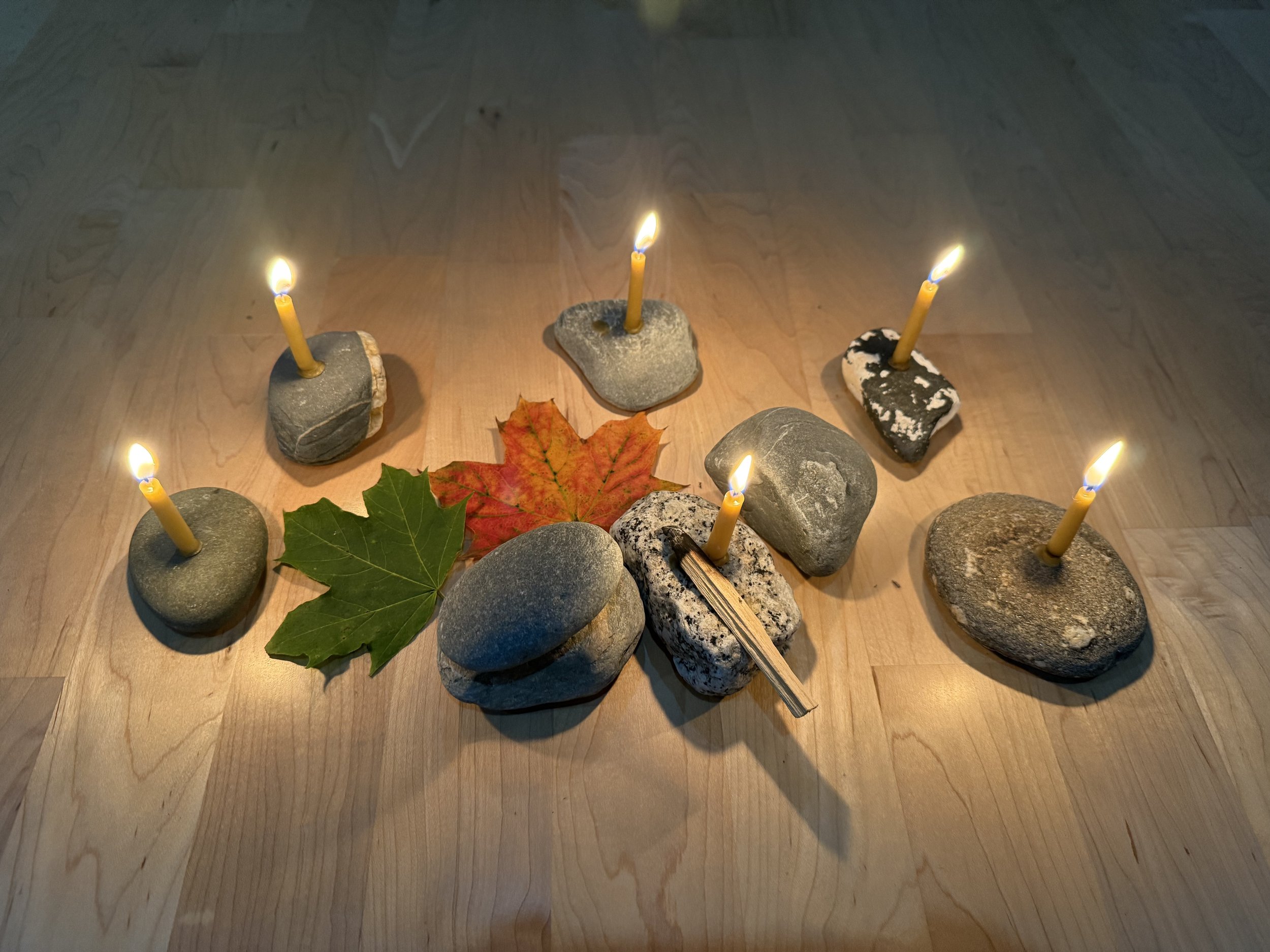 Five small lit candles placed in rocks on a wooden floor, with a maple leaf and two stones in the center.