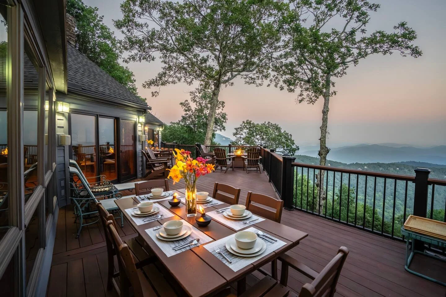A decorated outdoor wooden deck dining area overlooking a scenic mountain view during sunset, with a table set for six with bowls, plates, cutlery, a vase of flowers, and candles, surrounded by trees and a house.