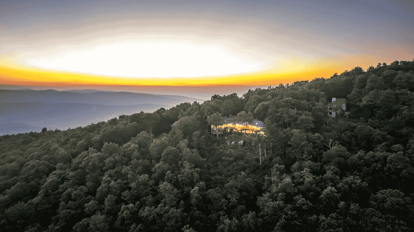 A mountain landscape at sunset with a house on the hillside illuminated by interior lights, surrounded by dense trees and distant mountain ranges.