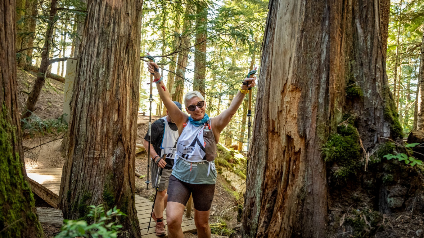 A smiling woman in sunglasses and athletic gear holding trekking poles in a forest during a trail race.