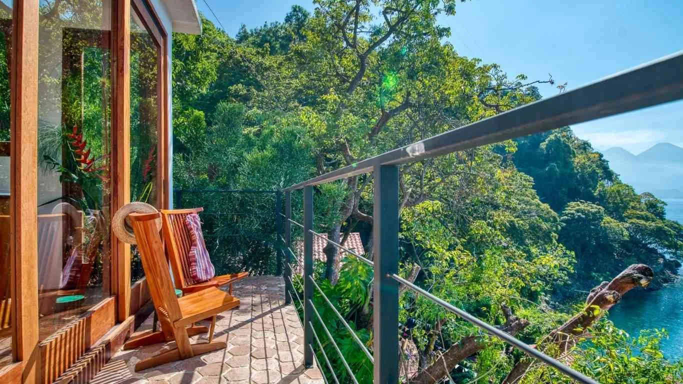 Balcony with wooden chairs overlooking lush green trees and blue water in the distance.