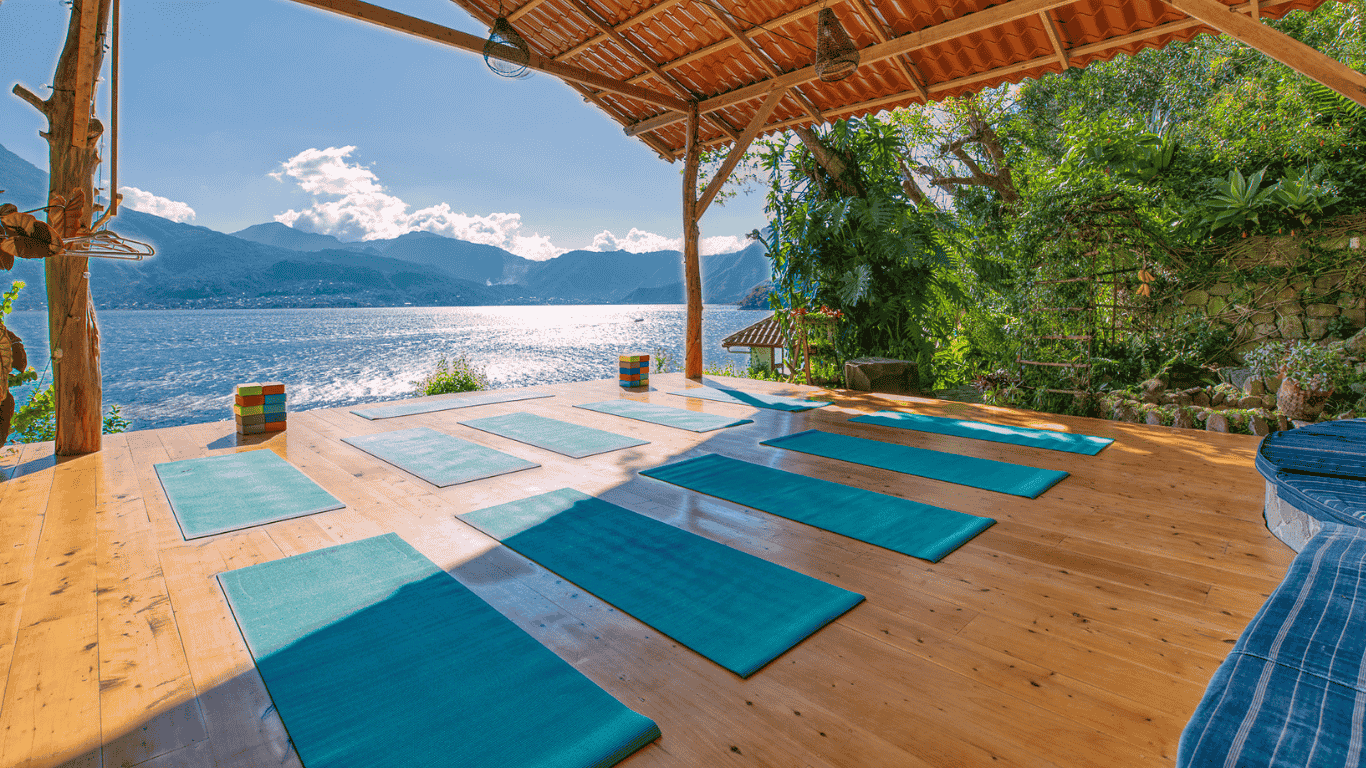 Yoga mats arranged on a wooden floor near a lake with mountains in the background, under a covered outdoor deck with a roof made of wood and clay tiles, surrounded by greenery.