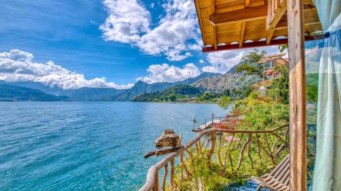 View from a cottage balcony overlooking a lake, with mountains in the background, a clear sky with scattered clouds, and a small, decorative wooden carved animal on the balcony railing.