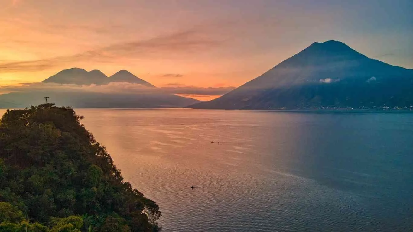 A scenic view of a lake with mountains in the background, including two volcanoes, during sunset with colorful sky.