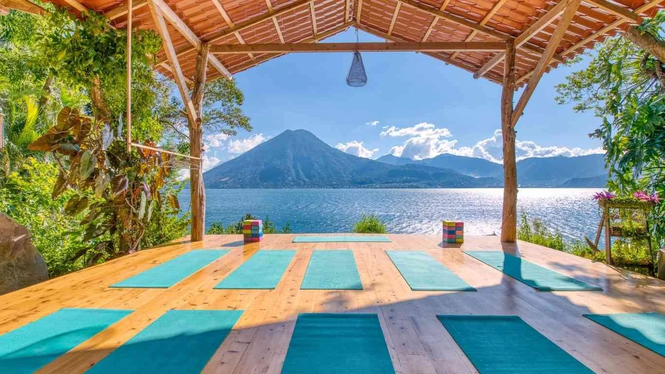 Open-air yoga studio with blue mats on wooden floor, overlooking a lake and volcanic mountain with partly cloudy sky in the background.