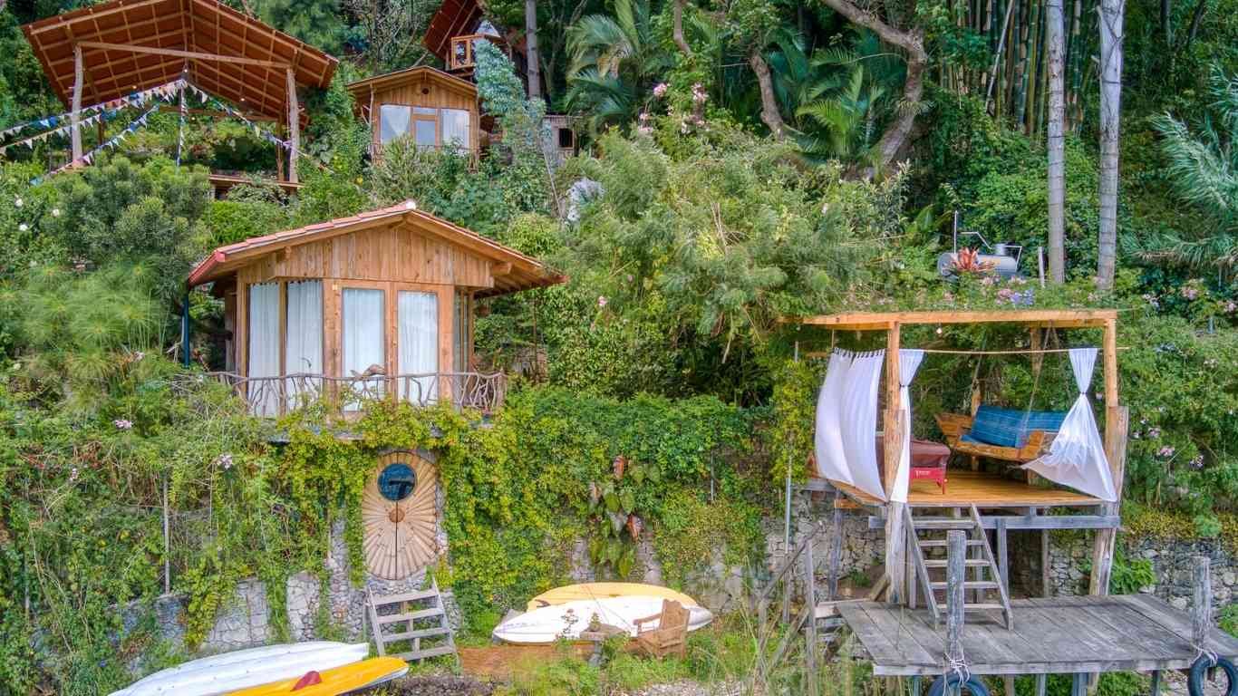 A hillside with multiple wooden treehouses and a wooden deck with a swing, surrounded by lush green trees and shrubs.