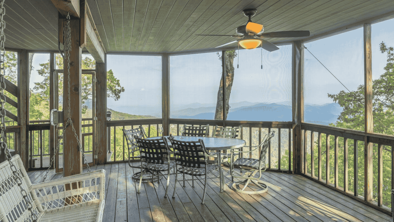Covered porch with wooden deck, round dining table with four chairs, ceiling fan, rocking swing, and scenic mountain view with trees.