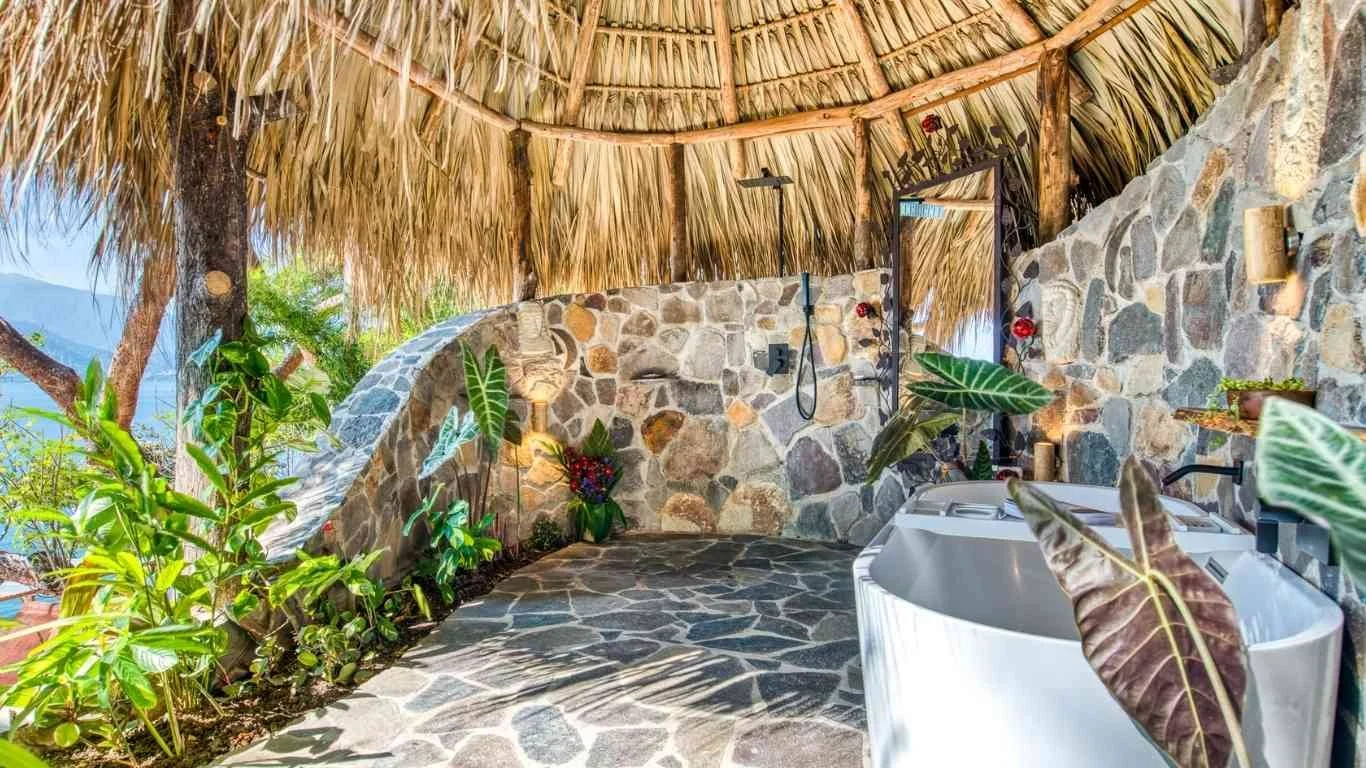 An outdoor shower area with a thatched roof and stone walls, decorated with green plants and flowers, including a white bathtub.