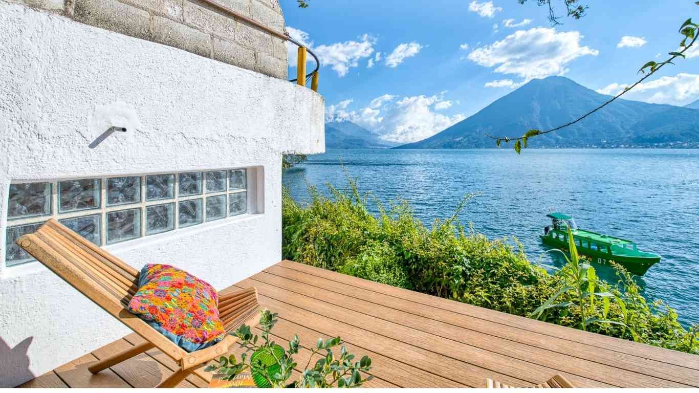 A wooden deck with a colorful pillow on a lounge chair, next to a white house with glass block windows, overlooking a lake and mountains with a green boat floating on the water.