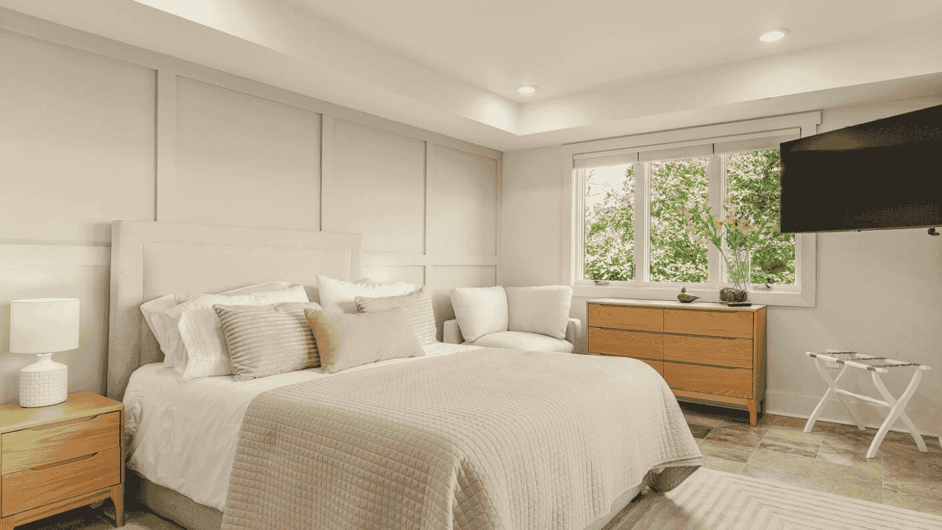Modern bedroom with a large bed, white headboard, beige bedding, and multiple pillows. There is a nightstand with a white lamp on the left, a wooden dresser near the window, and a wall-mounted flat-screen TV on the right. The room has large windows with greenery outside, and a small bench or tray in front of the TV.