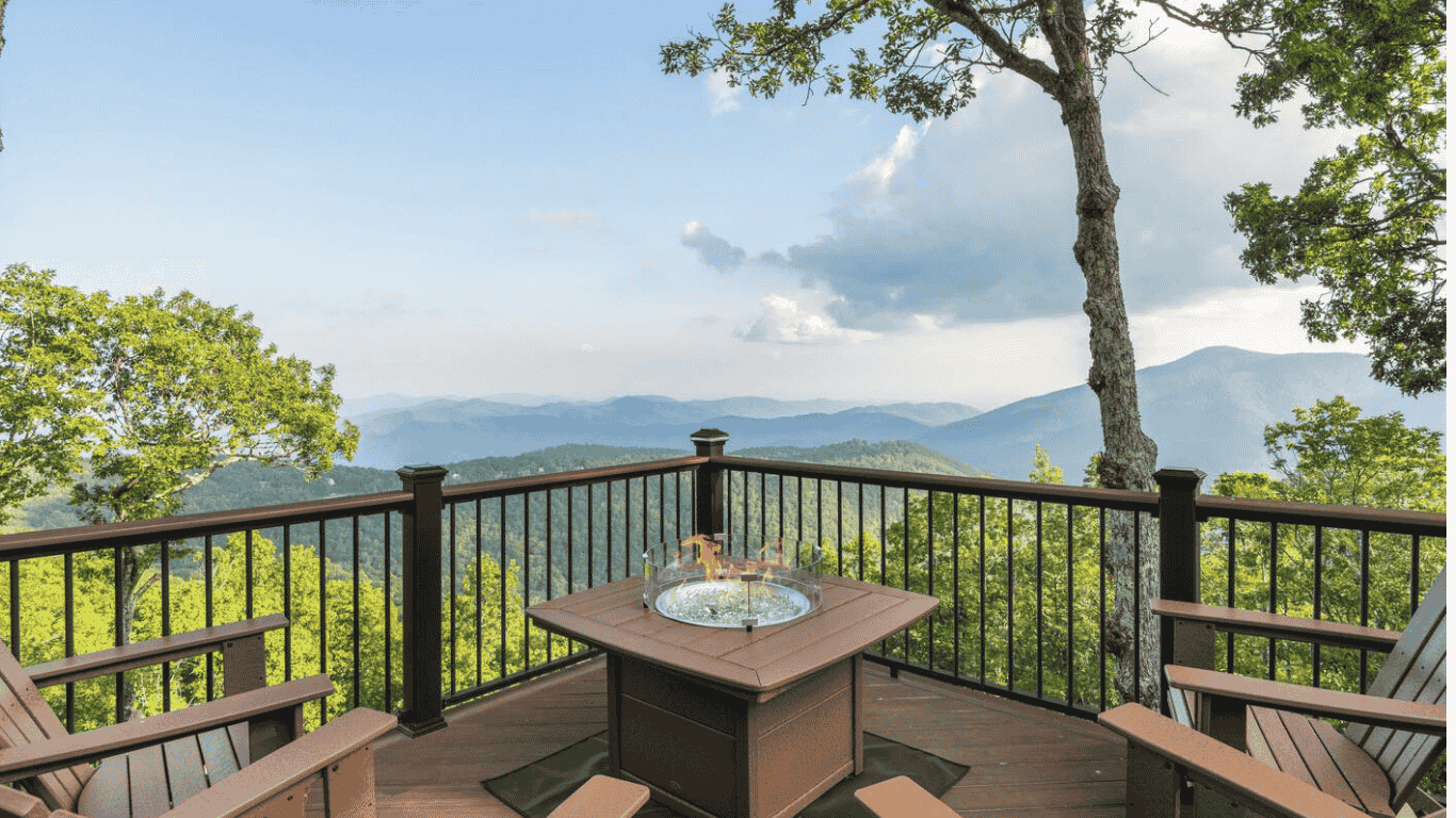 A wooden deck with patio chairs, overlooking a mountain landscape with trees and blue sky.