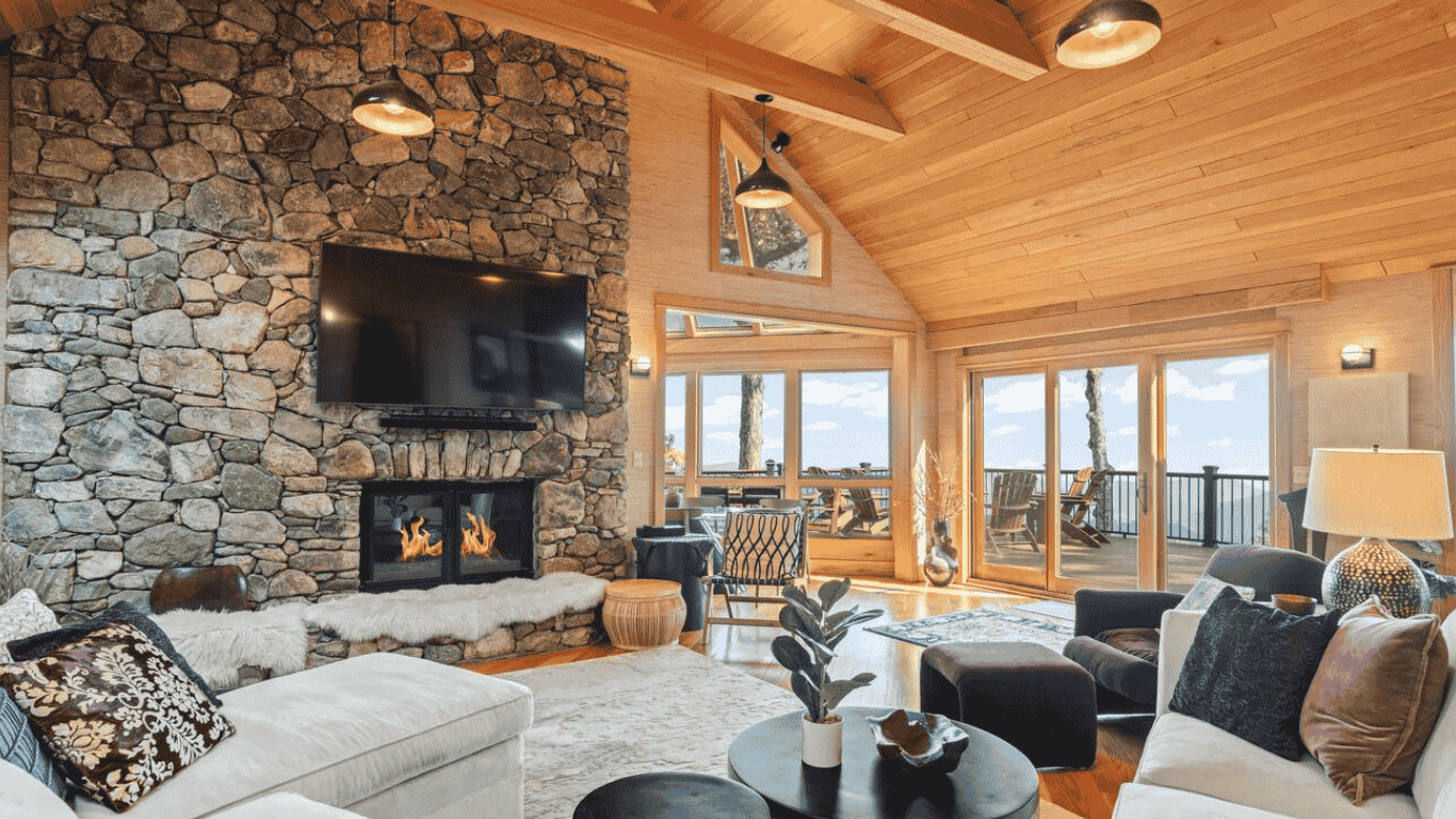 A cozy living room with wood-paneled vaulted ceiling, a stone fireplace with a mounted flat-screen TV, and large glass doors leading to a balcony with outdoor seating and a view of trees and water.