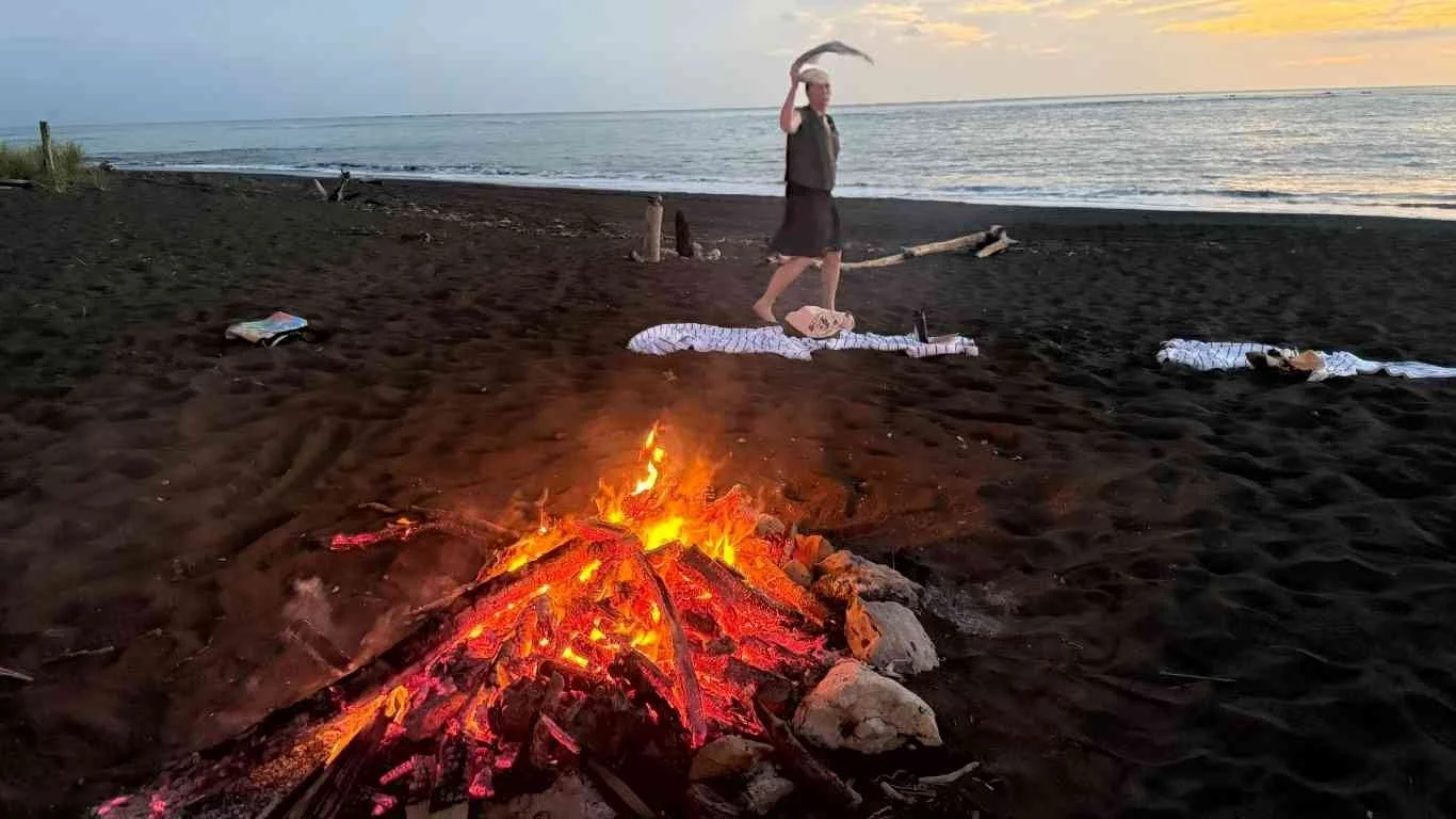Person dancing on a towel on the beach at sunset, with a campfire burning in the foreground.