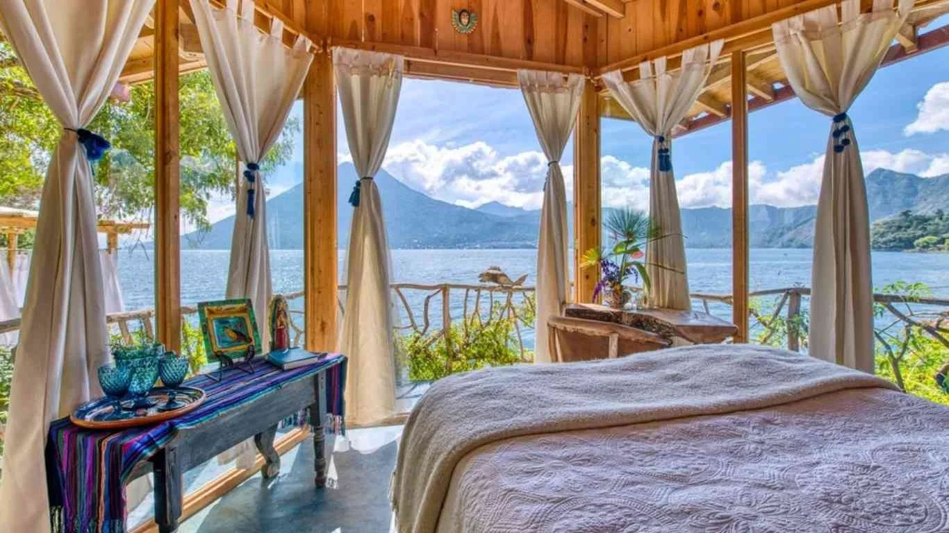 A bedroom with a view of a lake and mountains through large open windows with white curtains tied back. The bed has a cream-colored quilt. There is a wooden table with decorative blue glassware, a small framed artwork, and books. A small wooden table with a plant is near the window.