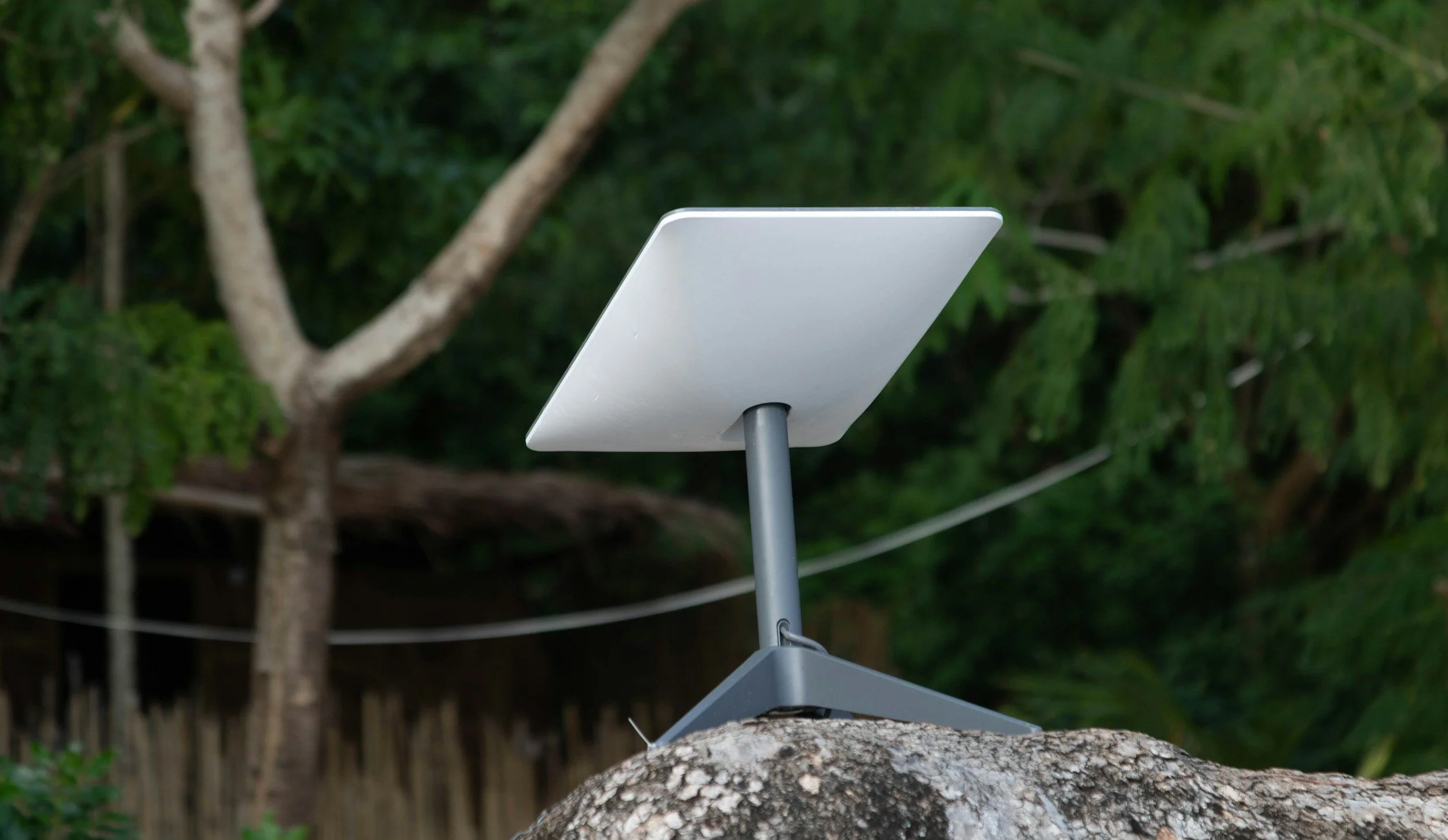 A weather station with a white rectangular sensor mounted on a stand, situated outdoors on a large rock, with green trees and a thatched roof structure in the background.