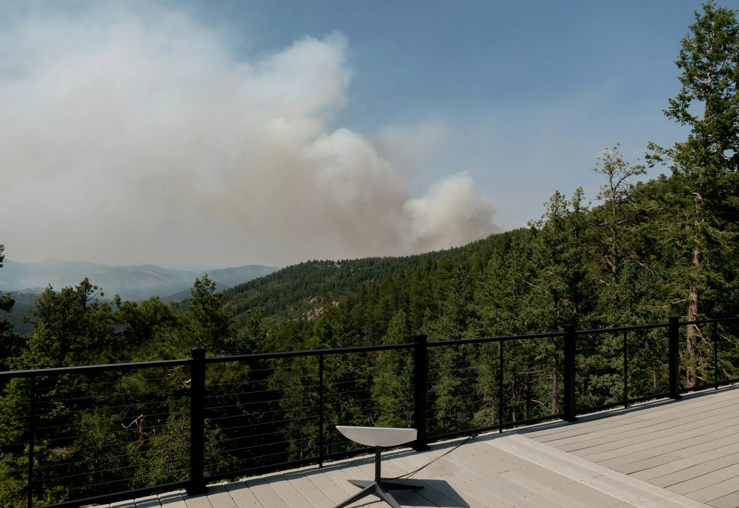 View from a balcony or rooftop overlooking a forested mountain landscape with smoke rising in the distance, possibly from a wildfire.