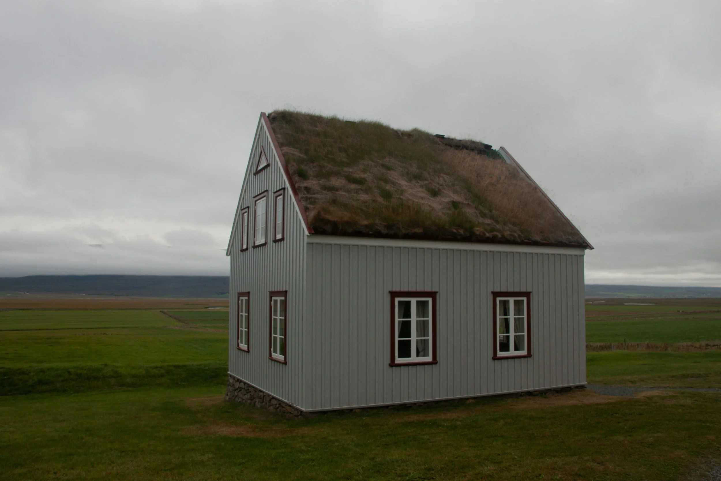 A house with a grass-covered roof and vertical gray siding, situated on a grassy field with open overcast sky in the background.
