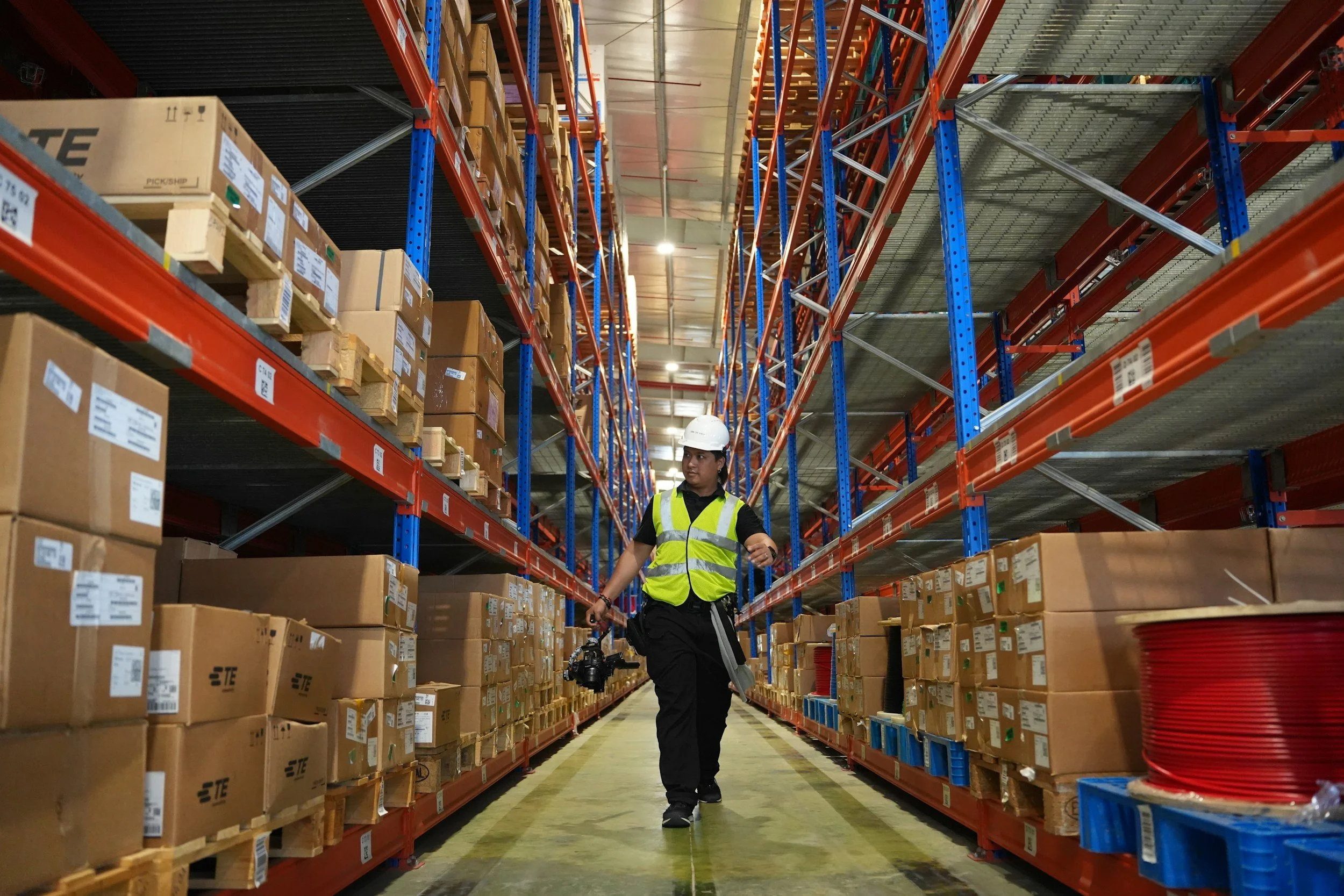 A person wearing a white safety helmet and high-visibility yellow vest walking through a warehouse aisle, surrounded by shelves filled with boxes and pallets.