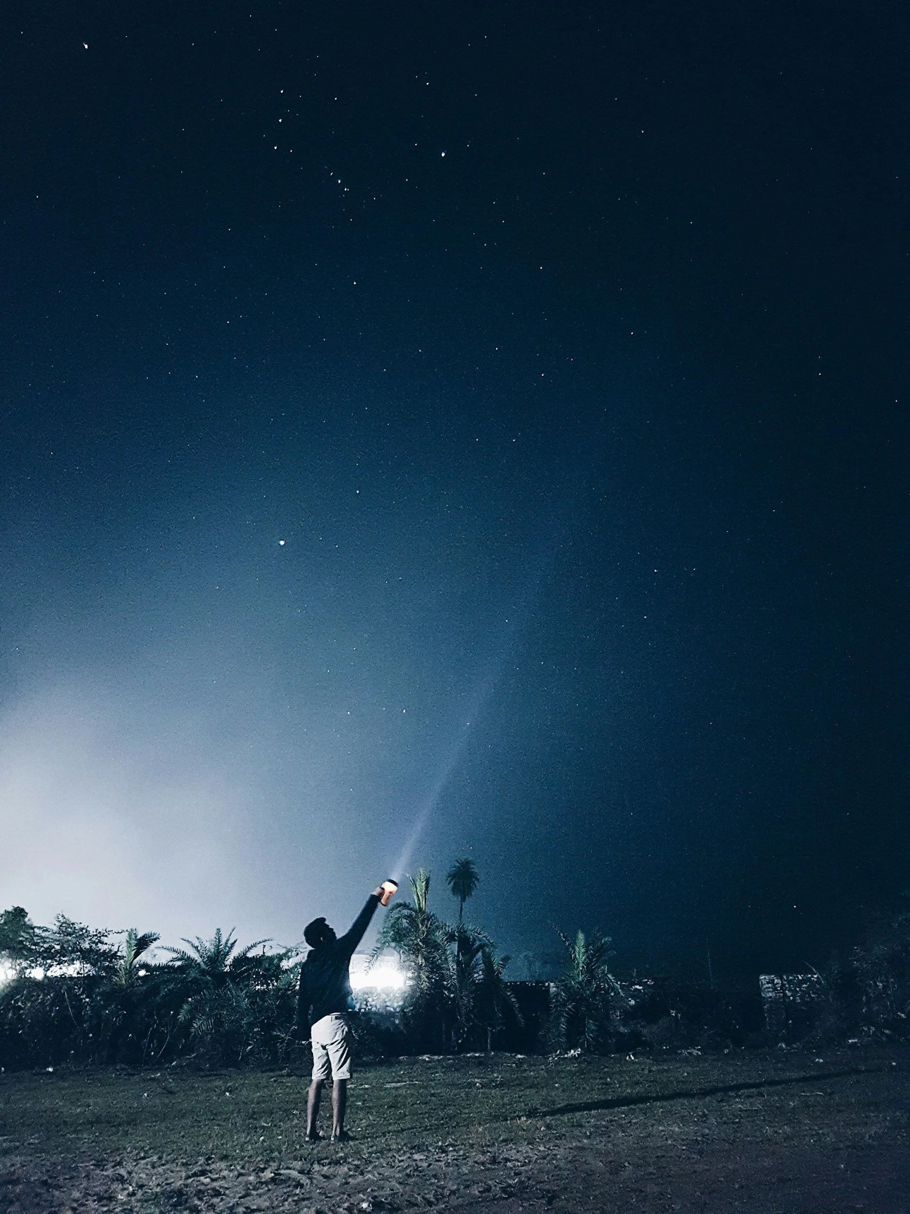 A person standing outdoors at night, holding a flashlight directed upward toward a star-filled sky with some visible clouds and palm trees in the background.