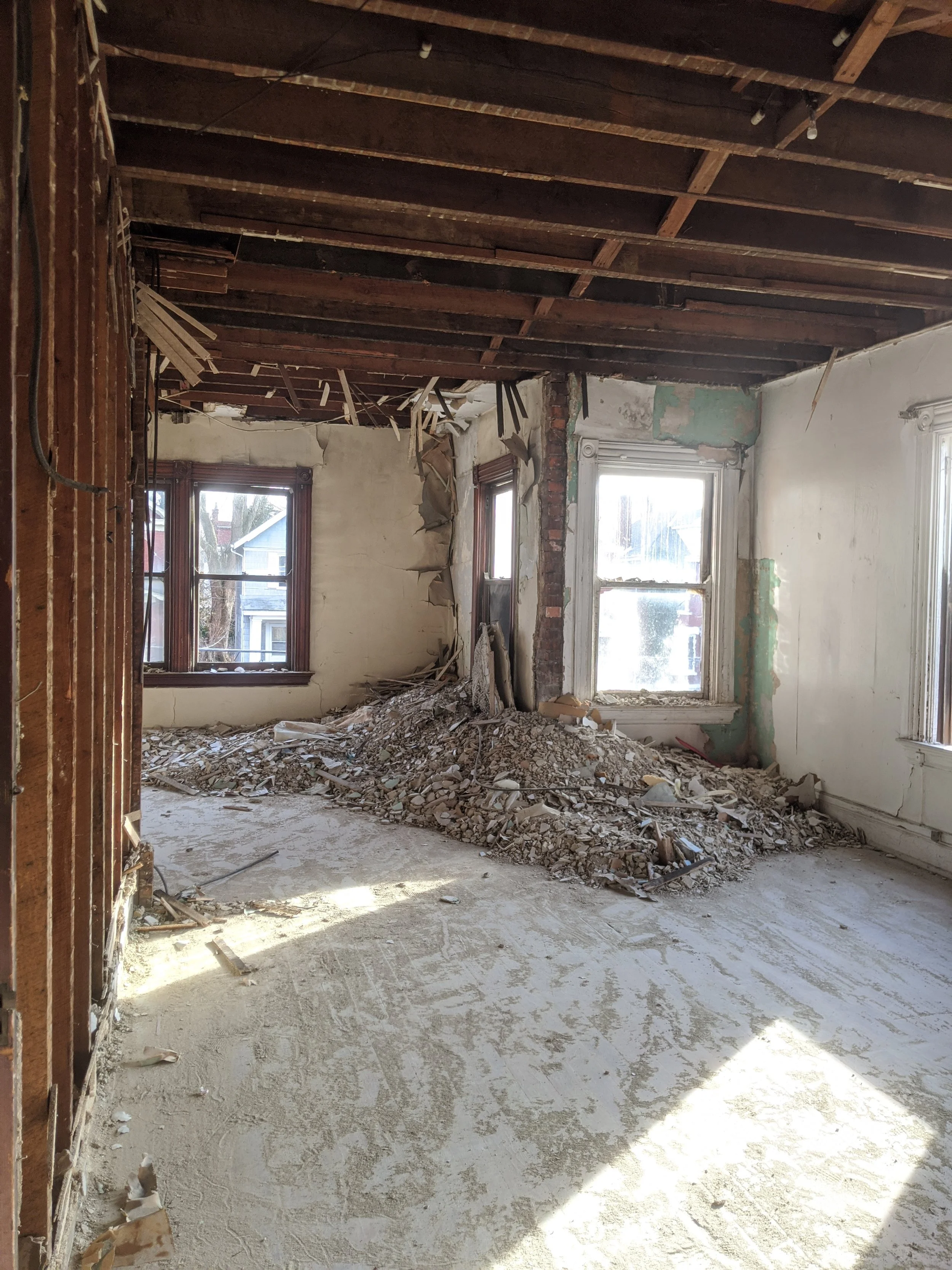 Room under demolition with exposed ceiling beams, broken walls, and debris piled in the center, sunlight coming through windows.