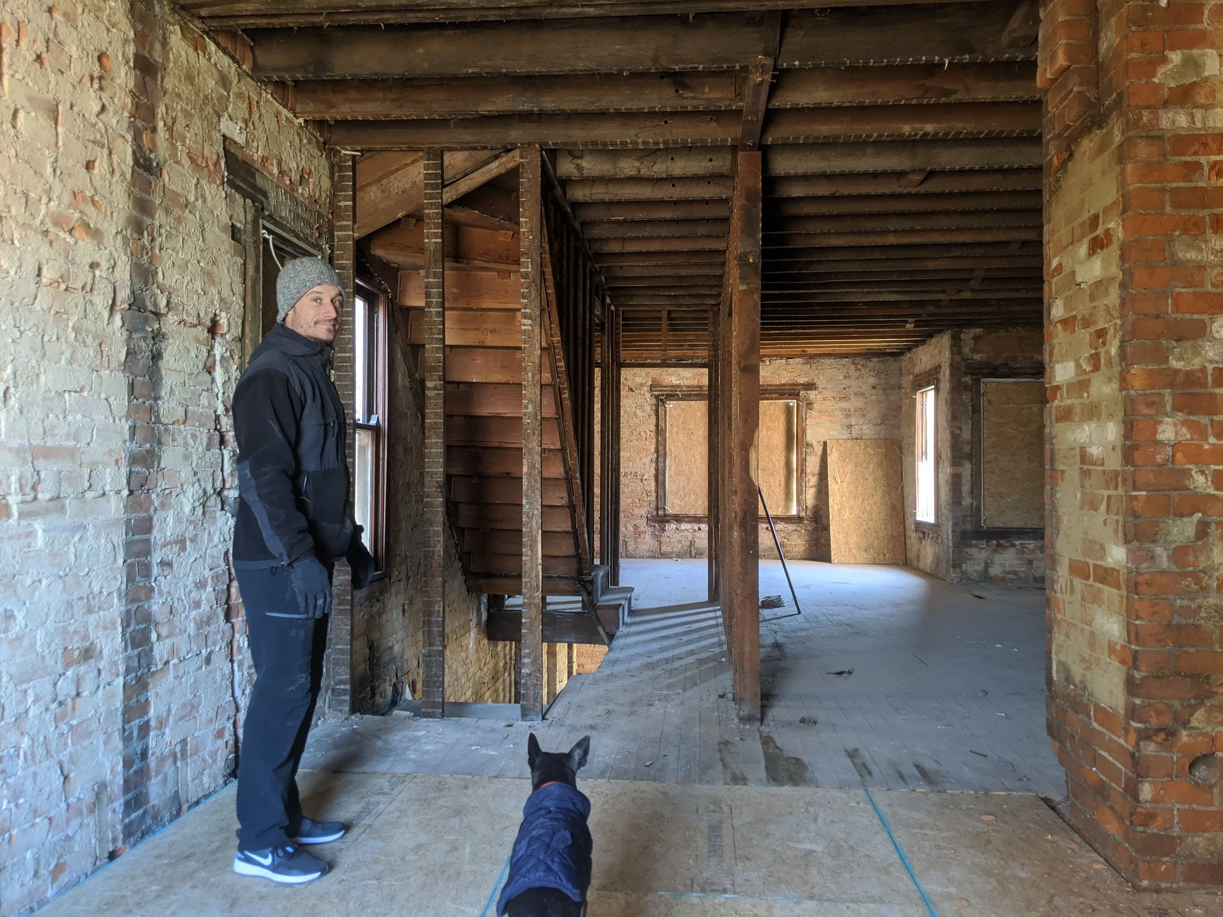A man wearing a gray beanie, black jacket, and gloves stands inside an unfinished brick and wood building. A dog wearing a blue vest is in the foreground, facing toward the man. The interior features exposed brick walls, wooden ceilings, and an open staircase.