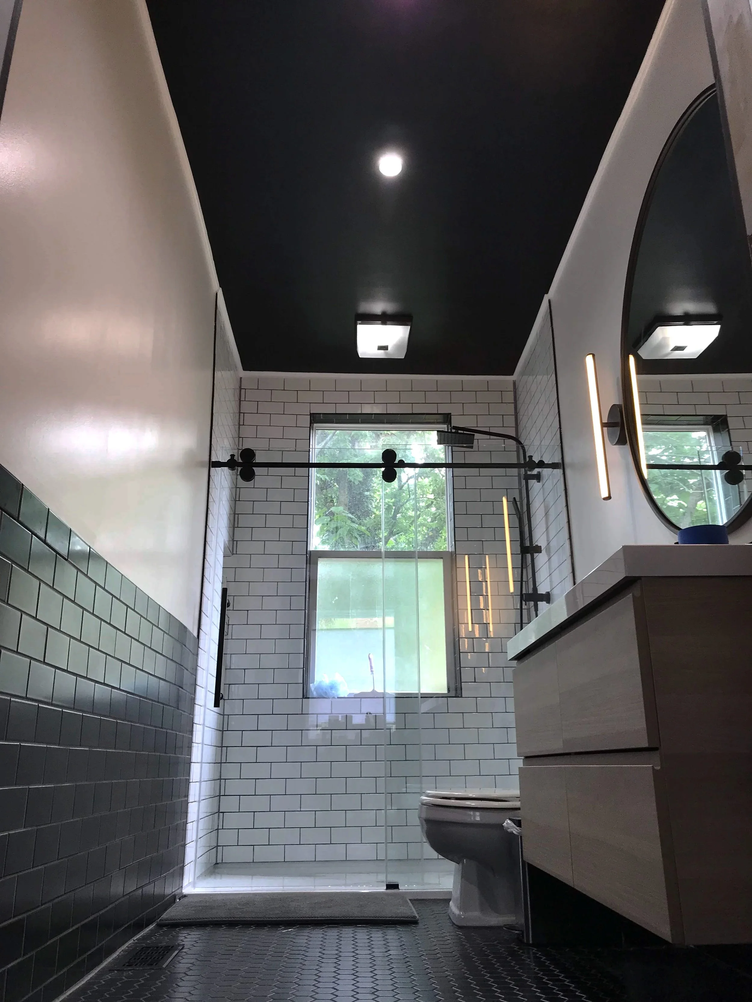 Modern bathroom with black ceiling, white subway-tile walls, a glass shower enclosure, a window with green trees outside, a floating vanity with a round mirror, and a toilet.