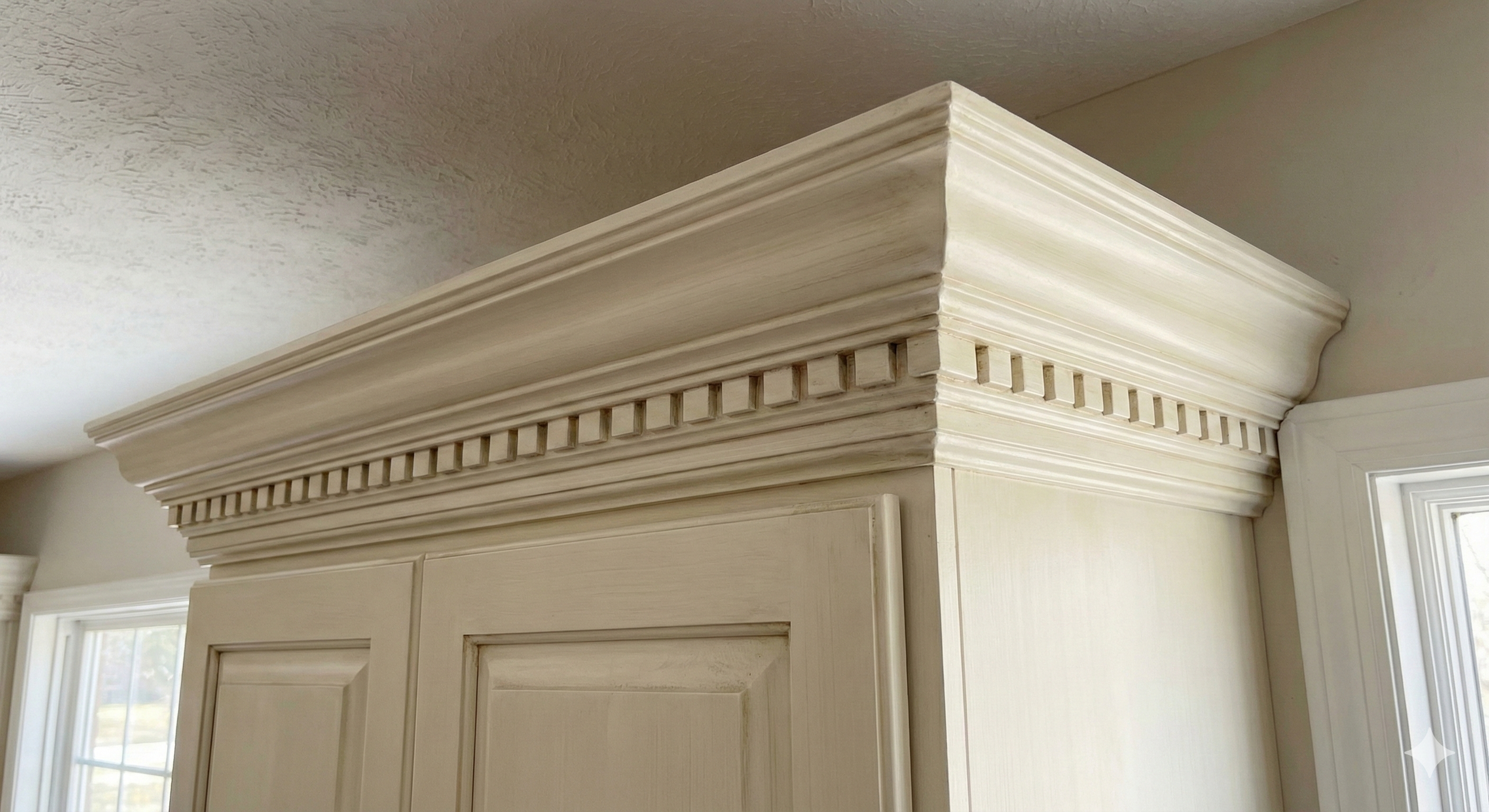 Close-up of decorative crown molding in a light cream color above a cabinet, with a textured ceiling and a window nearby.