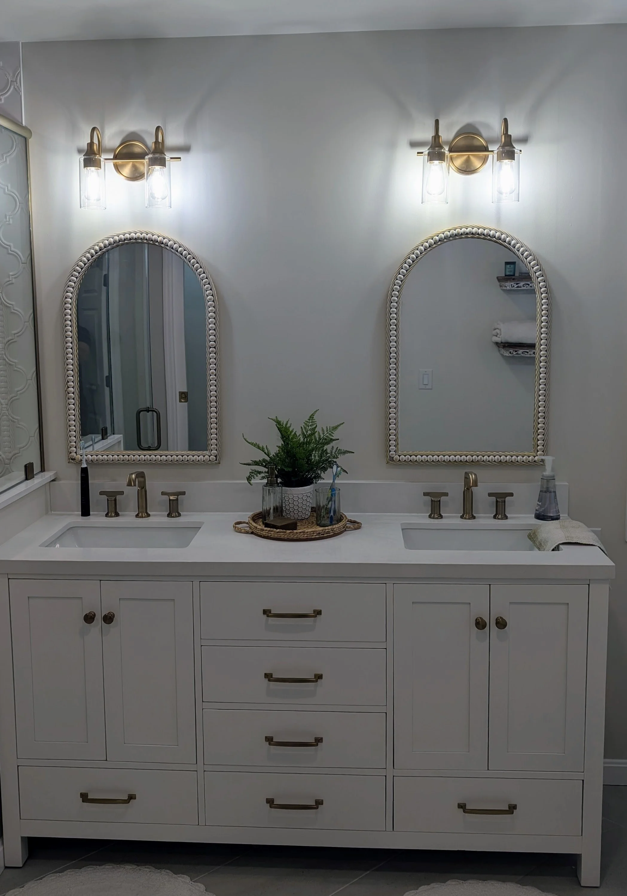 Bathroom vanity with two sinks, two mirrors with beaded frames, brass fixtures, and a potted plant on a tray in the center. There are wall-mounted light fixtures above each mirror and shelves with towels on the right.