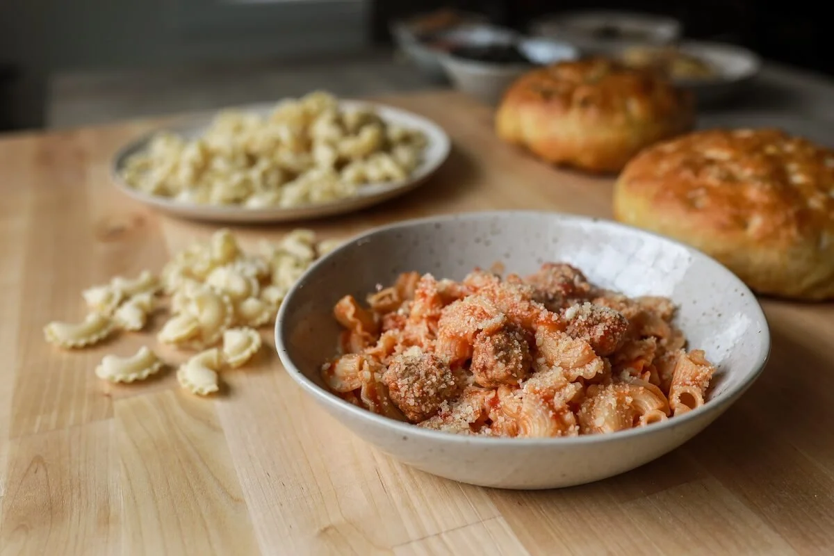 Bowls of pasta with tomato sauce and grated cheese, garlic bread rolls, and a plate of cauliflower on a wooden table.