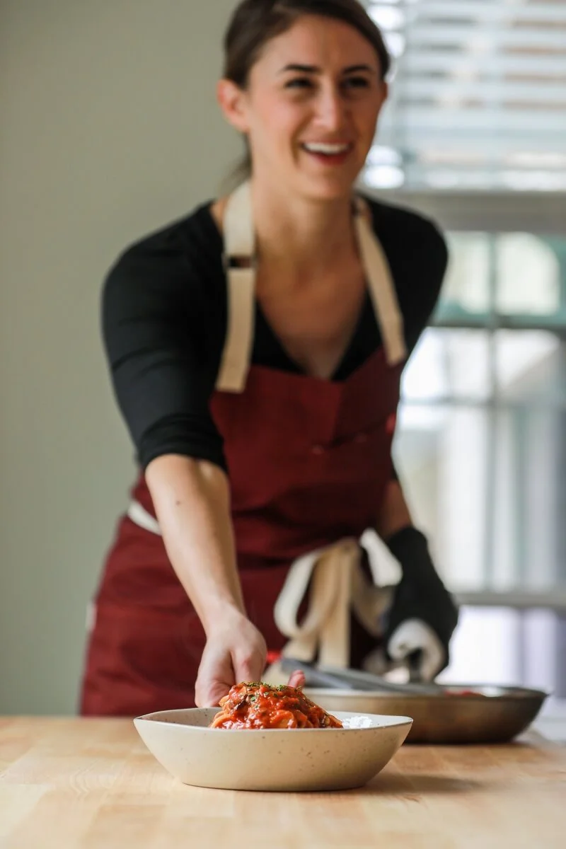 Woman in a red apron serving food onto a plate in a bright kitchen.