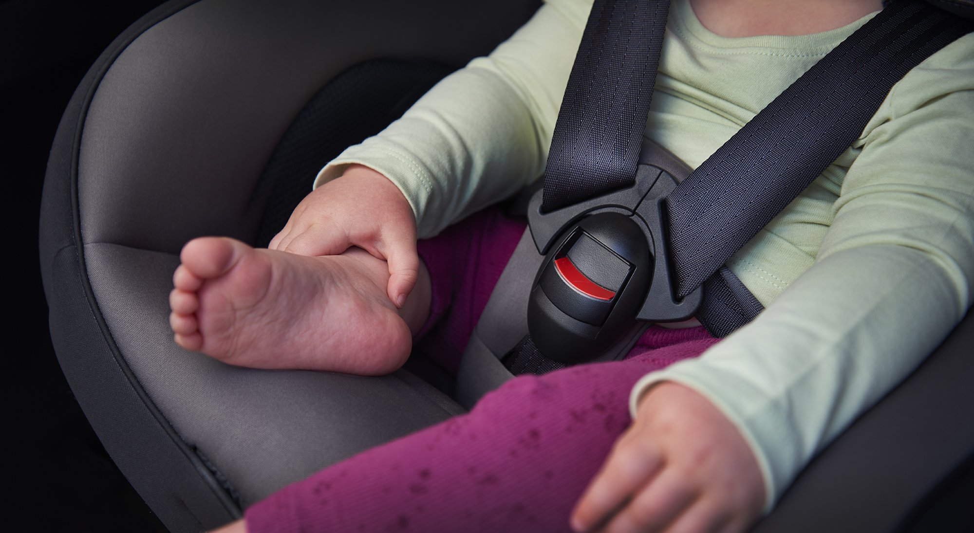 Close-up of a child's foot in a car seat with a five-point harness, with hands resting on their lap.