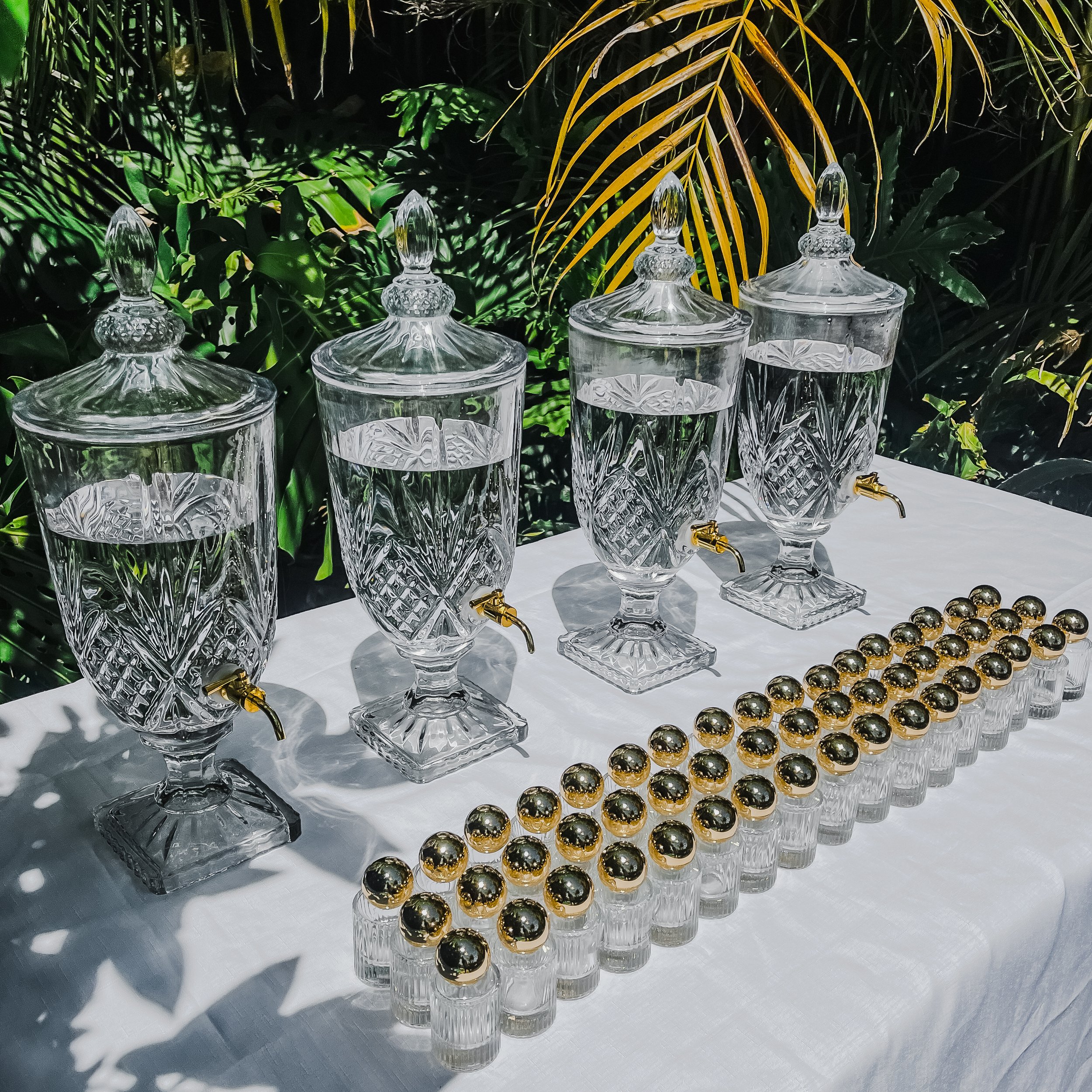 Four glass perfume dispensers with gold spigots on a white tablecloth, against lush green tropical foliage background, with three rows of perfume bottles with round gold lids