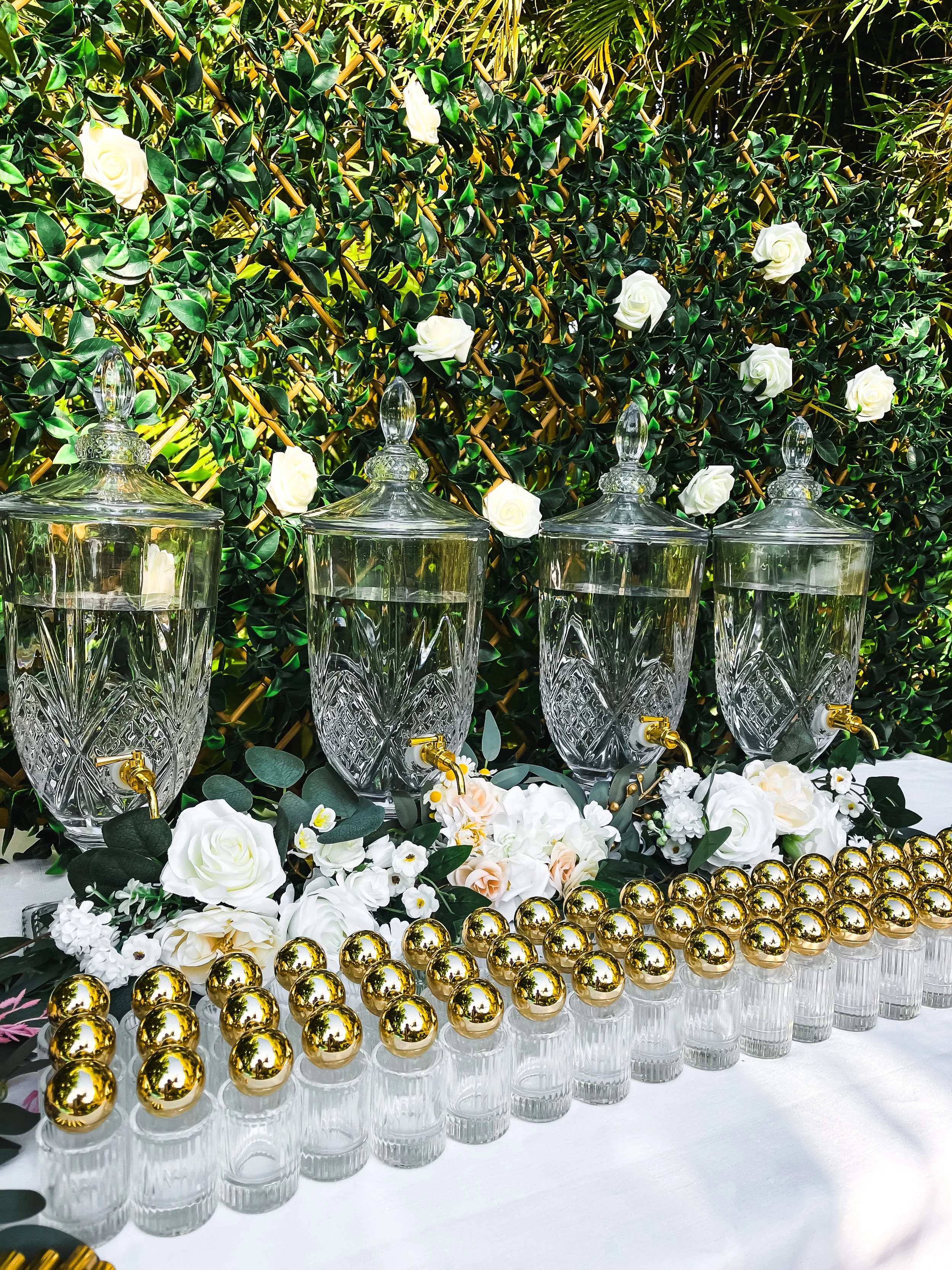 Elegant outdoor table setup with four glass perfume dispensers with gold spigots, surrounded by white and cream flowers and a lush green leafy backdrop, with three rows of perfume bottles with round gold lids.