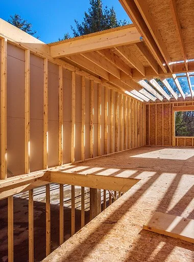 Interior view of a building under construction with exposed wooden framing, sunlight casting shadows, and a partially finished roof with transparent panels.